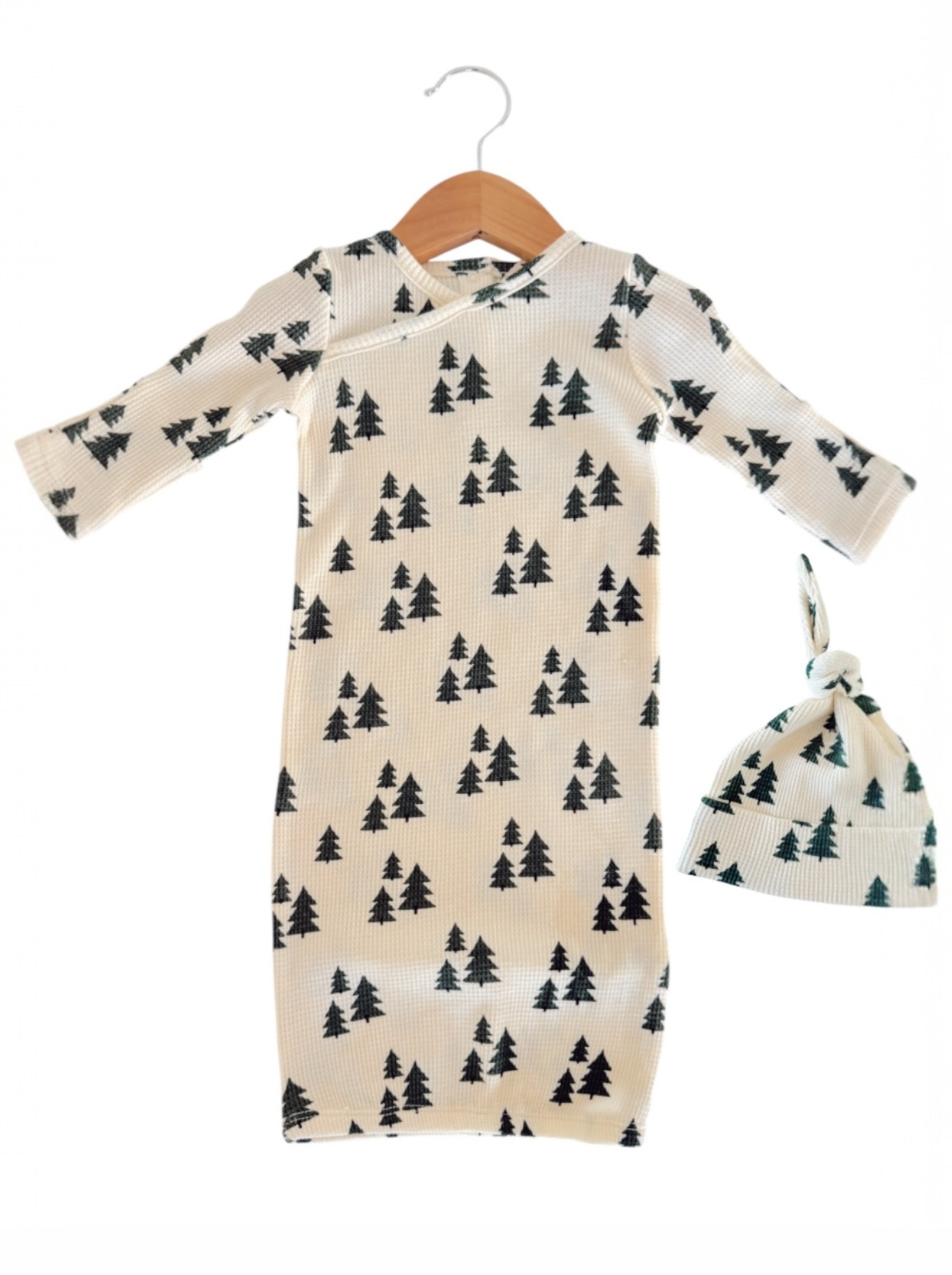 Cream-colored baby gown with black tree patterns, paired with a matching knotted hat, hanging on a wooden hook.
