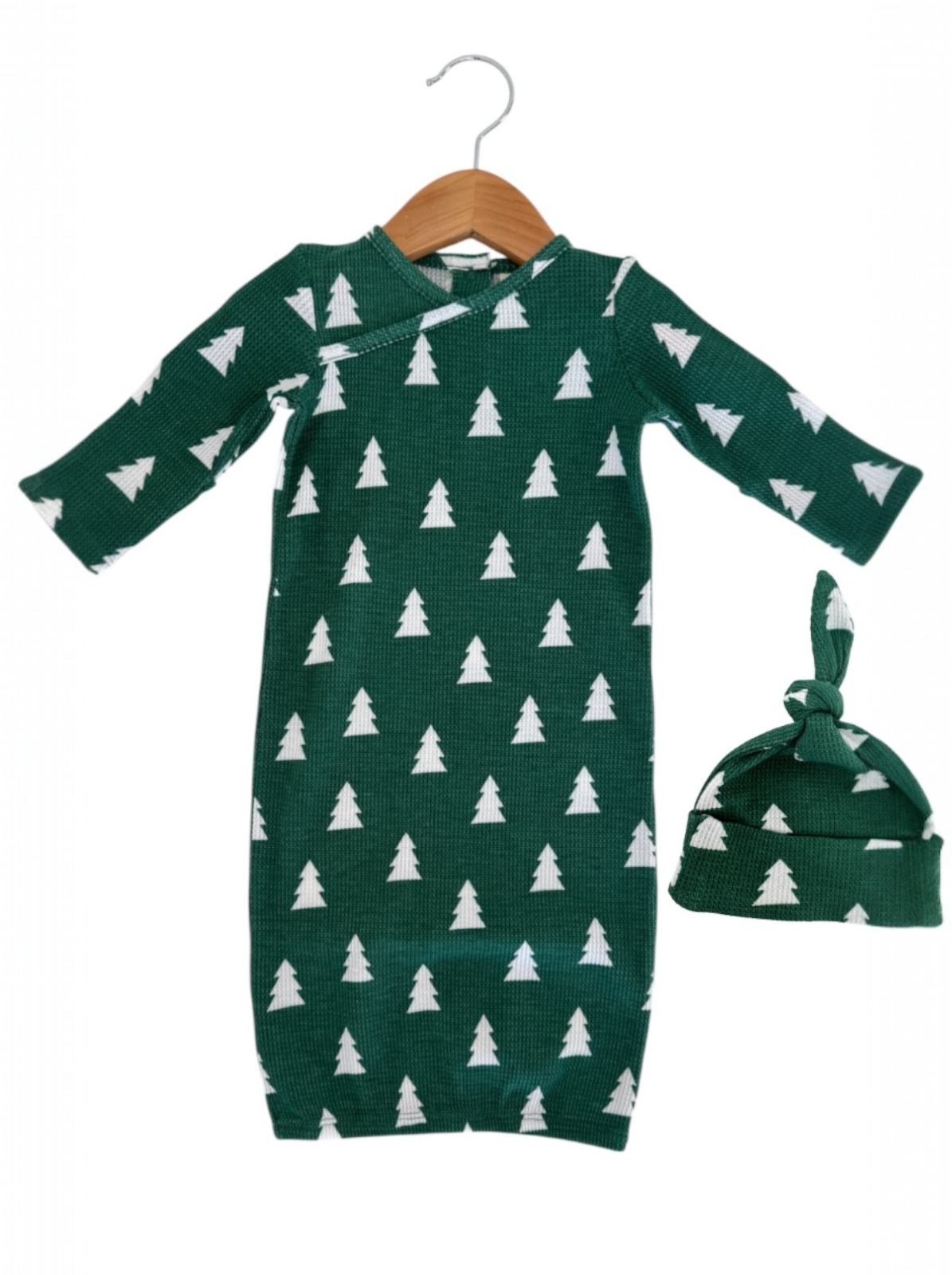 Green baby gown with white tree patterns, paired with a matching knotted hat, hanging on a wooden hanger.