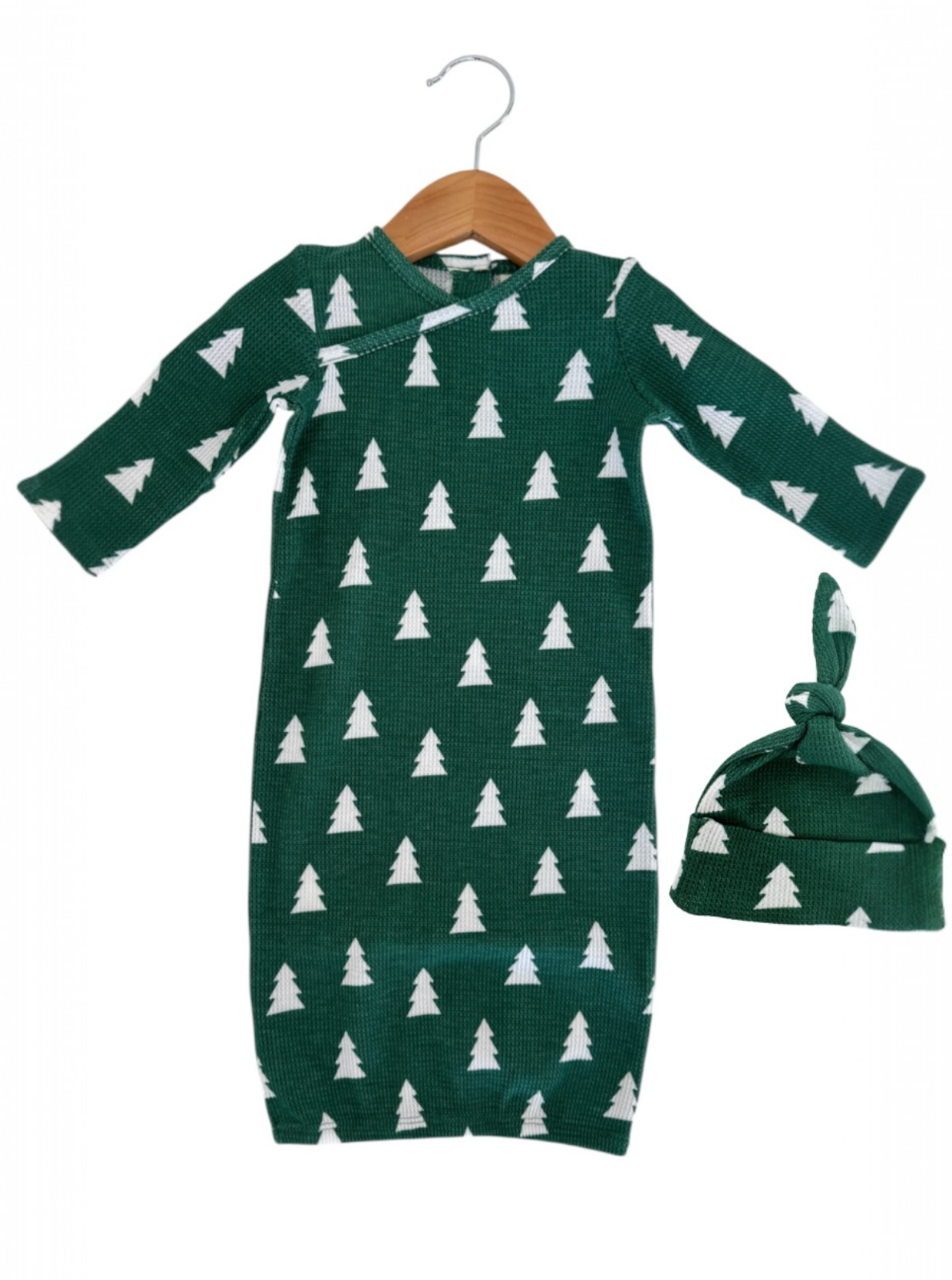 Green baby gown with white tree patterns, paired with a matching knotted hat, hanging on a wooden hanger.