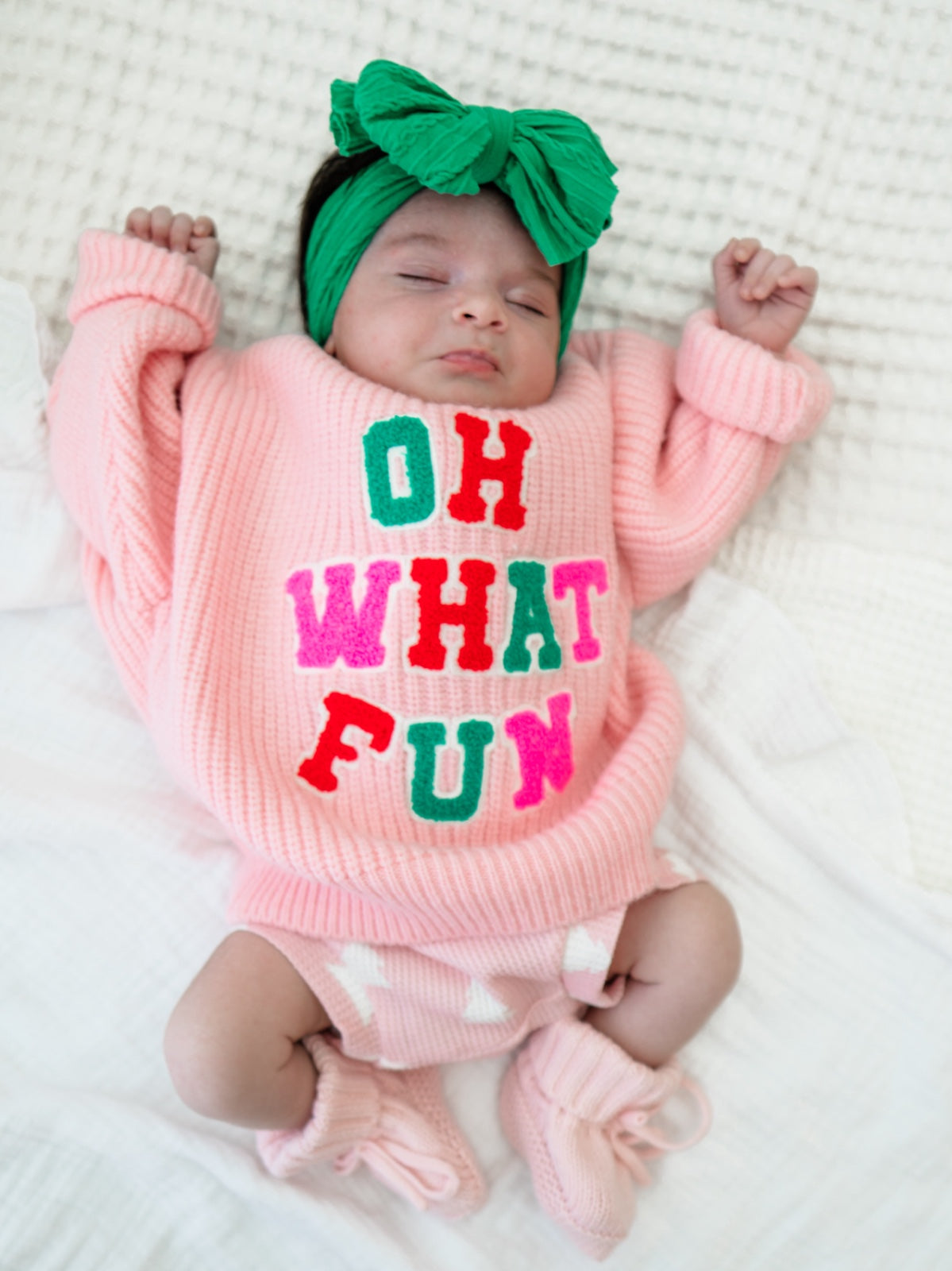 Infant wearing a pink sweater with "OH WHAT FUN" text and a green headband, lying on a white blanket.