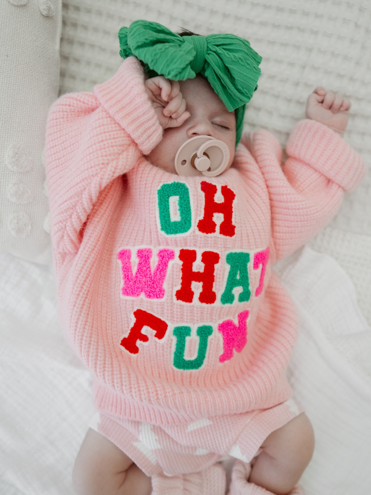 Baby in a pink sweater with colorful letters "OH WHAT FUN," wearing a green bow, resting peacefully on a white blanket.