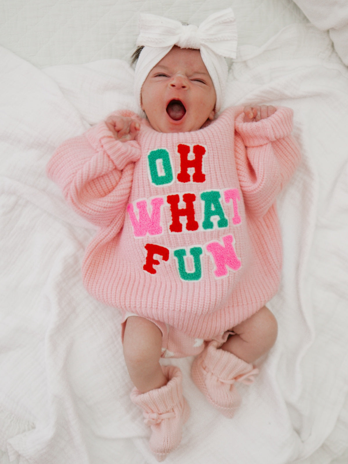 Baby yawning in a pink sweater with "OH WHAT FUN" text, wearing a bow headband and cozy booties.