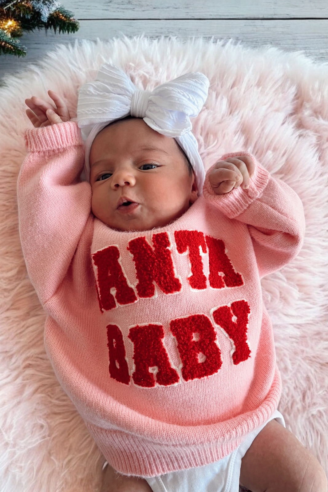 Baby girl in a pink sweater with "Santa Baby" text, wearing a white bow, lying on a fluffy pink blanket.