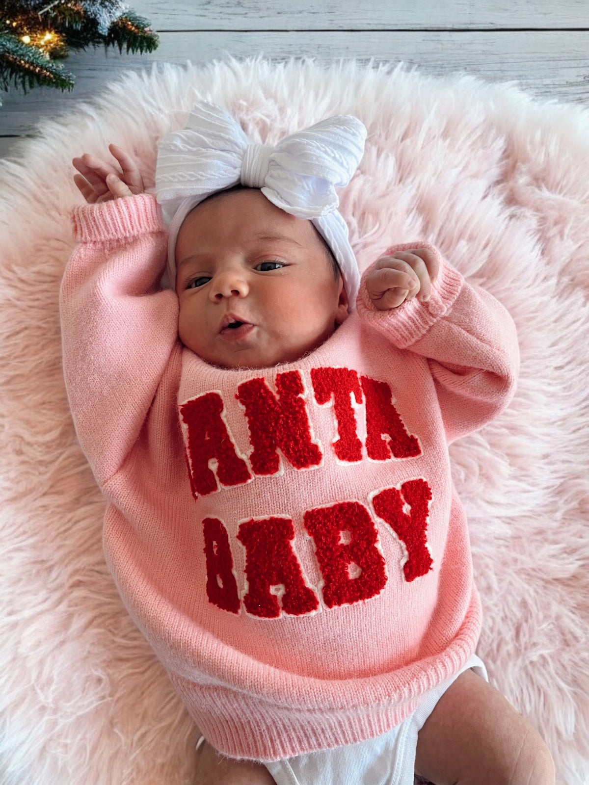 Baby girl in a pink sweater with "Santa Baby" text, wearing a white bow, lying on a fluffy pink blanket.