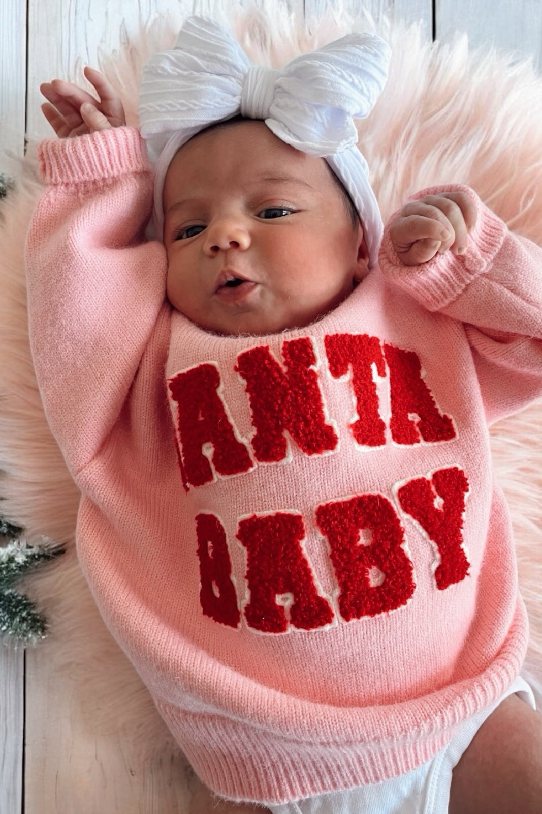 A baby in a pink sweater with "SANTA BABY" text and a white headband, lying on a fluffy pink surface.