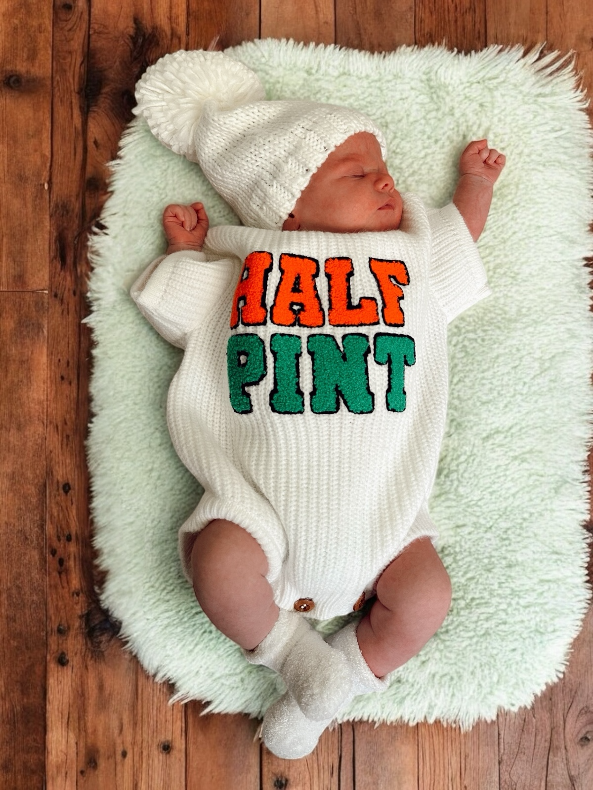 Baby wearing a white outfit with "HALF PINT" in orange and green, lying on a fluffy mint green mat.