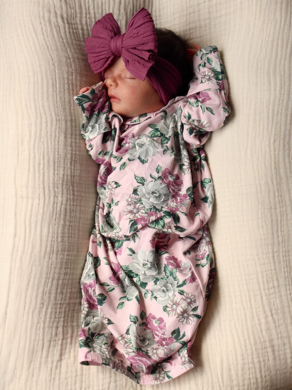 A sleeping baby in a floral pink outfit and a large purple headband on a soft textured blanket.