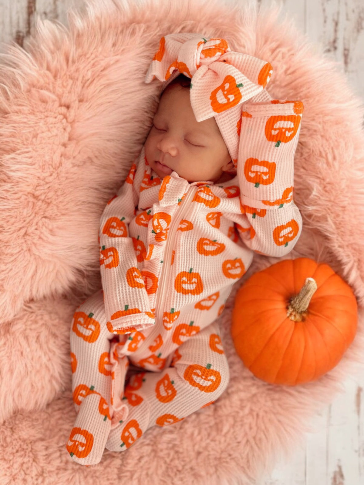 Sleeping baby in pumpkin-patterned outfit on pink fluff, next to a small orange pumpkin.