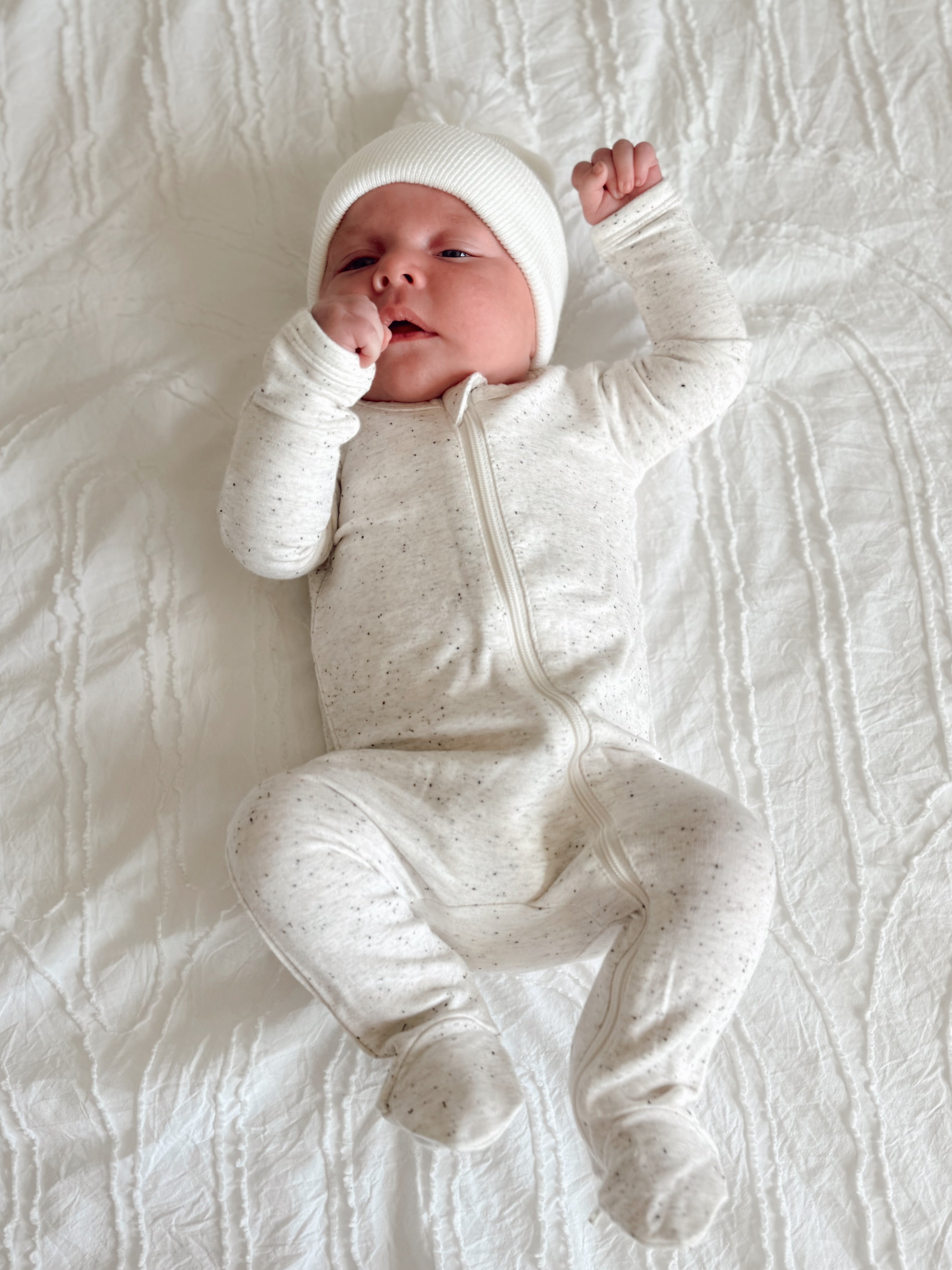Baby in a cozy cream onesie and white hat, laying on a textured white blanket, looking content.