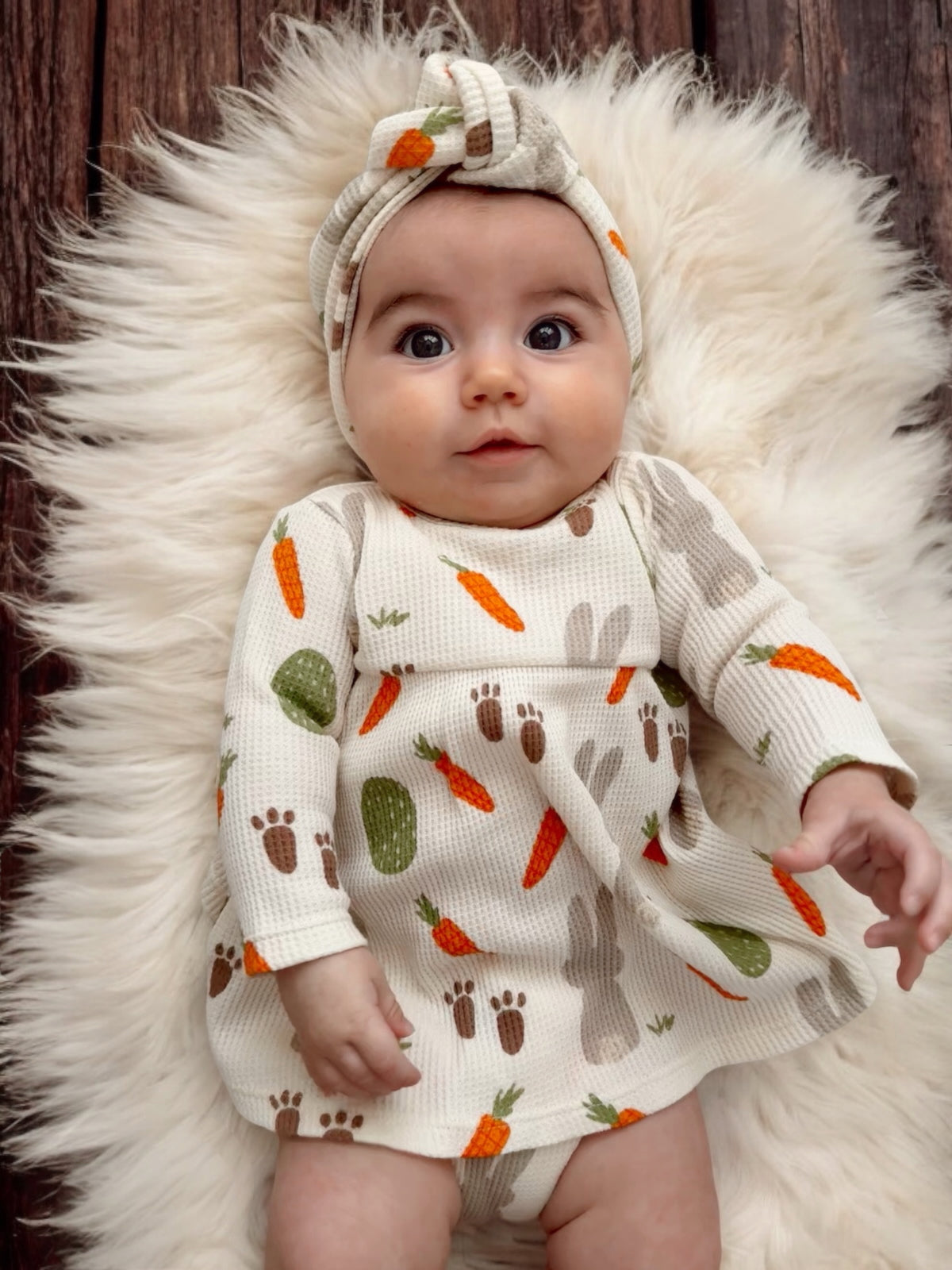 Baby girl in a patterned dress with carrots and bunnies, lying on a fluffy rug, smiling at the camera.