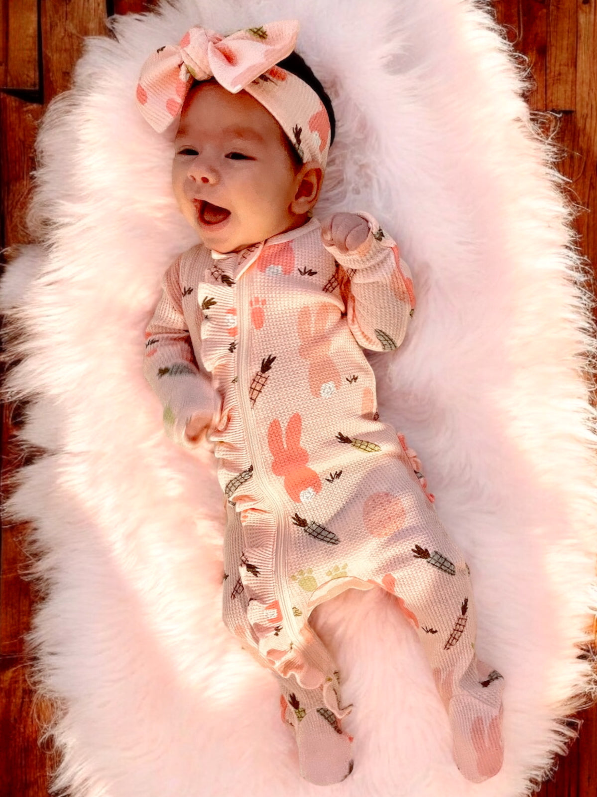 Smiling baby in pink outfit with bunny patterns, lying on a fluffy white blanket.