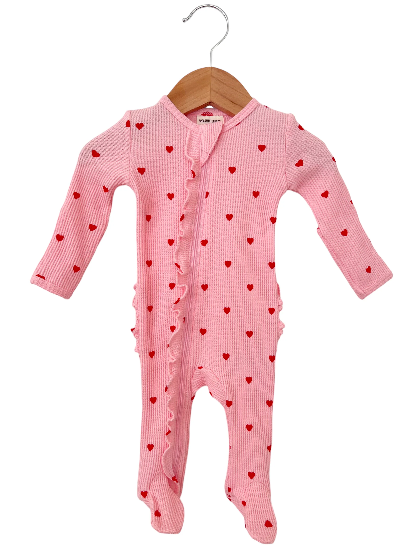 Pink baby onesie with red heart prints and ruffle detailing, hanging on a wooden hanger.