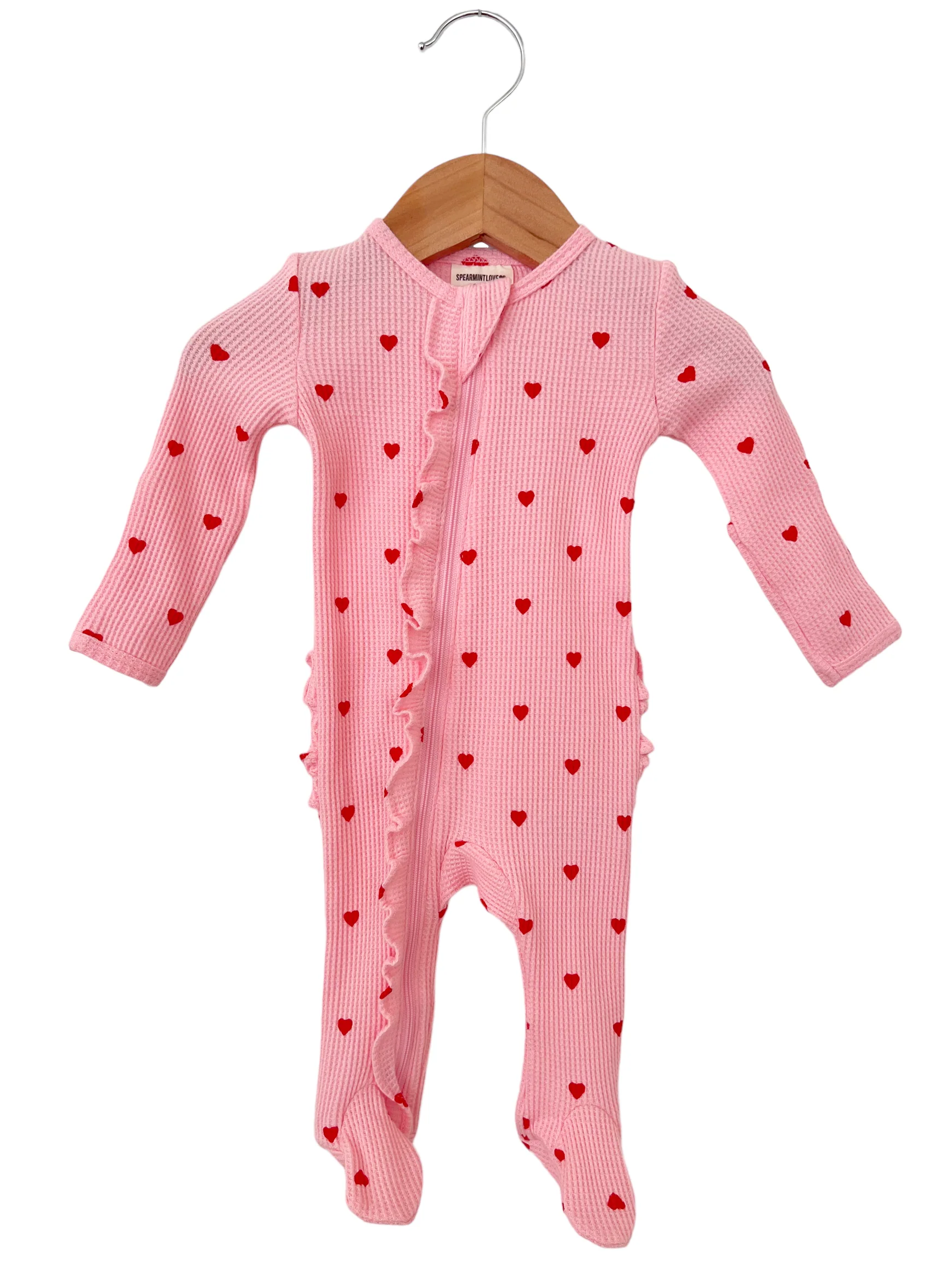Pink baby onesie with red heart prints and ruffle detailing, hanging on a wooden hanger.