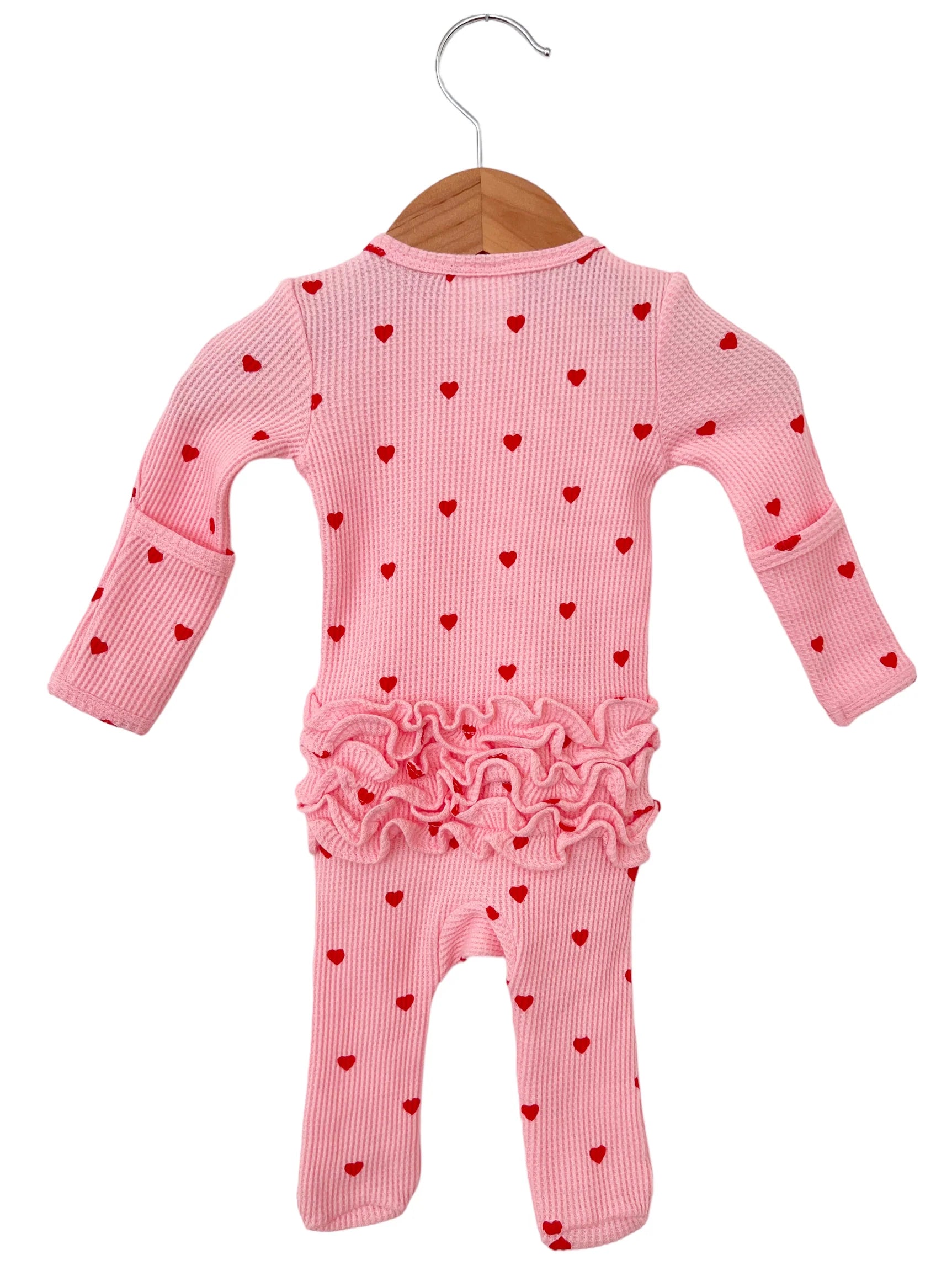 Pink baby onesie with red heart patterns and ruffled hem, hung on a wooden hanger.