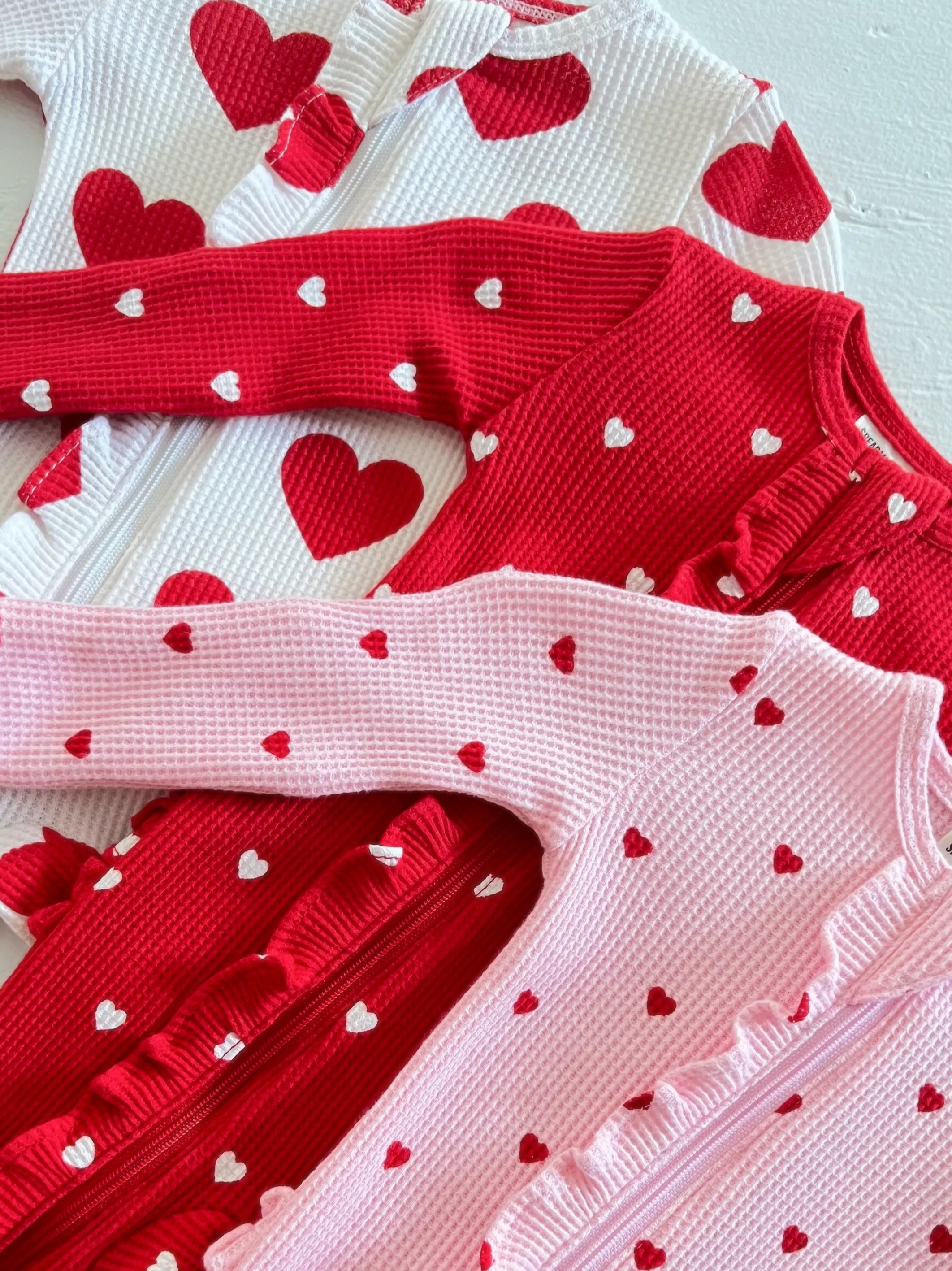 Three knitted cardigans in red, white, and pink, adorned with small heart patterns.