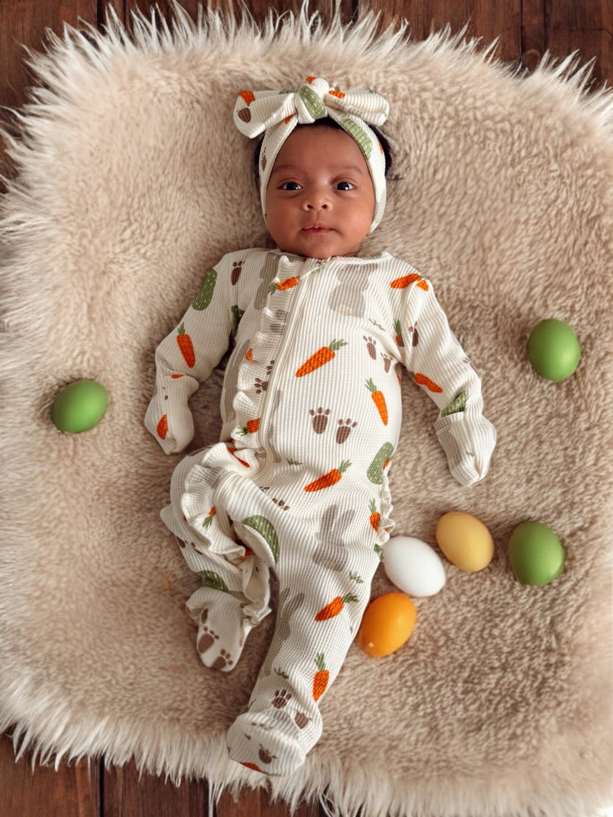 Baby in a cozy outfit with carrots and bunnies, surrounded by colorful eggs on a soft blanket.