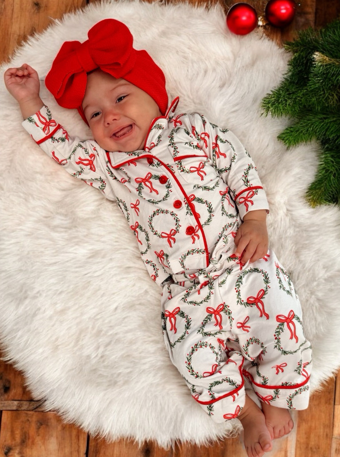 Smiling baby in festive pajamas with wreath and bow pattern, lying on a faux fur rug, with holiday decorations nearby.