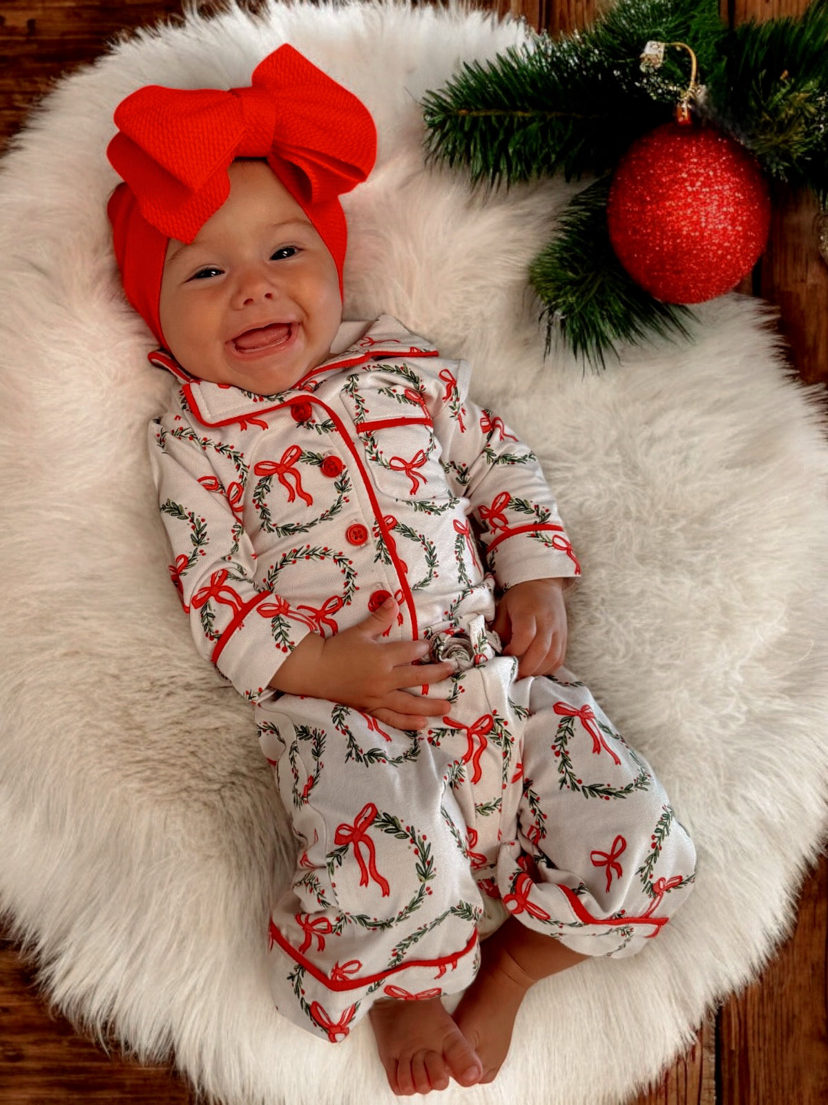Smiling baby in festive outfit with red bow, surrounded by holiday decorations on a cozy white blanket.