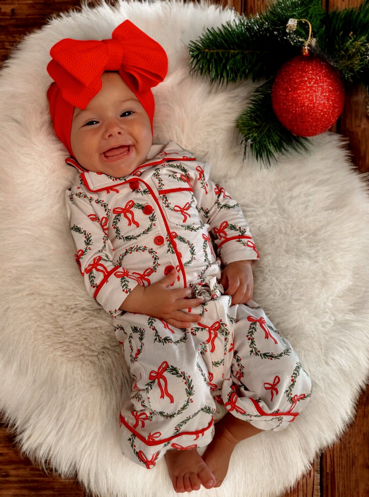 Smiling baby in festive outfit with red bow, surrounded by holiday decorations on a cozy white blanket.