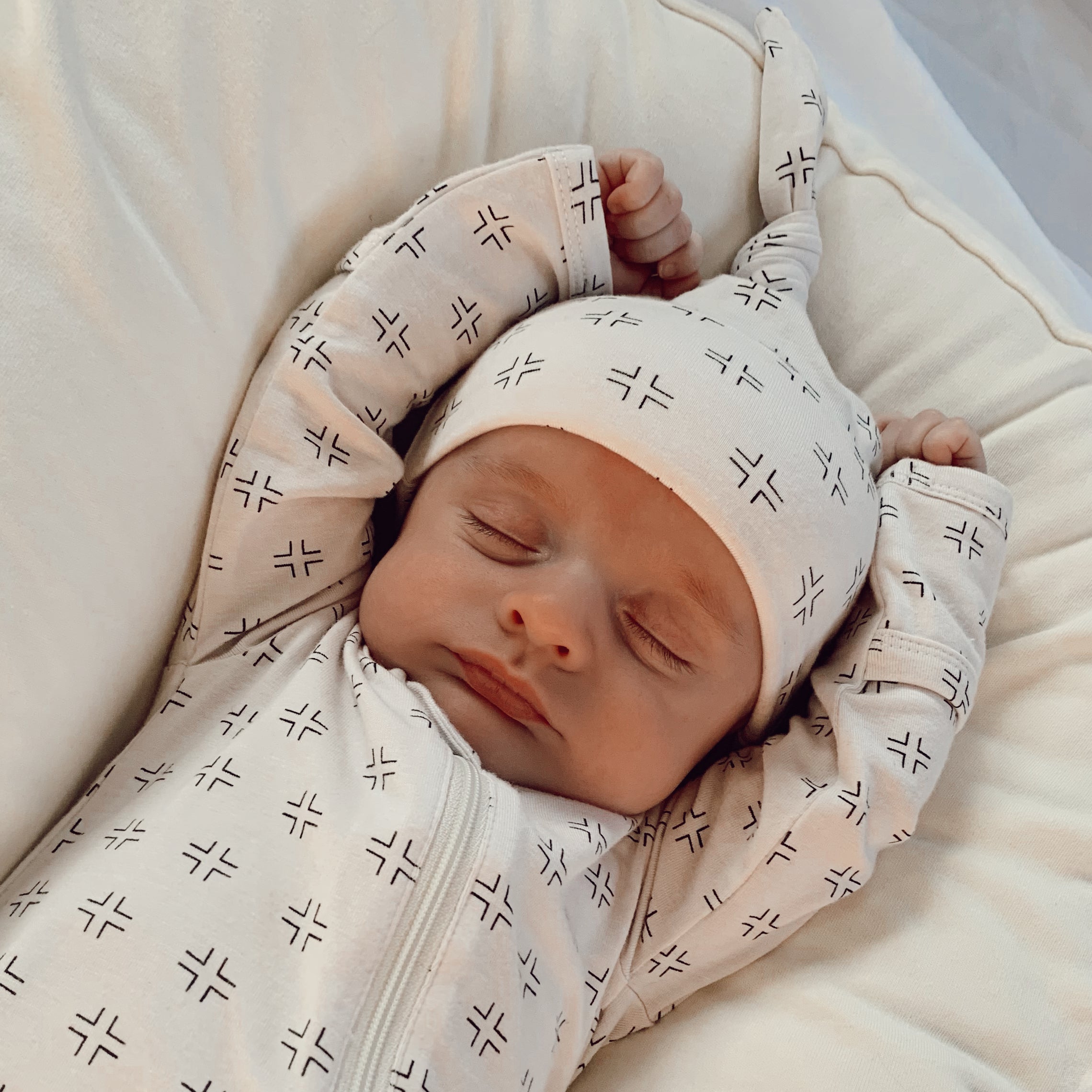 Sleeping baby in patterned pajamas, with arms raised and wearing a matching hat, resting on a soft surface.