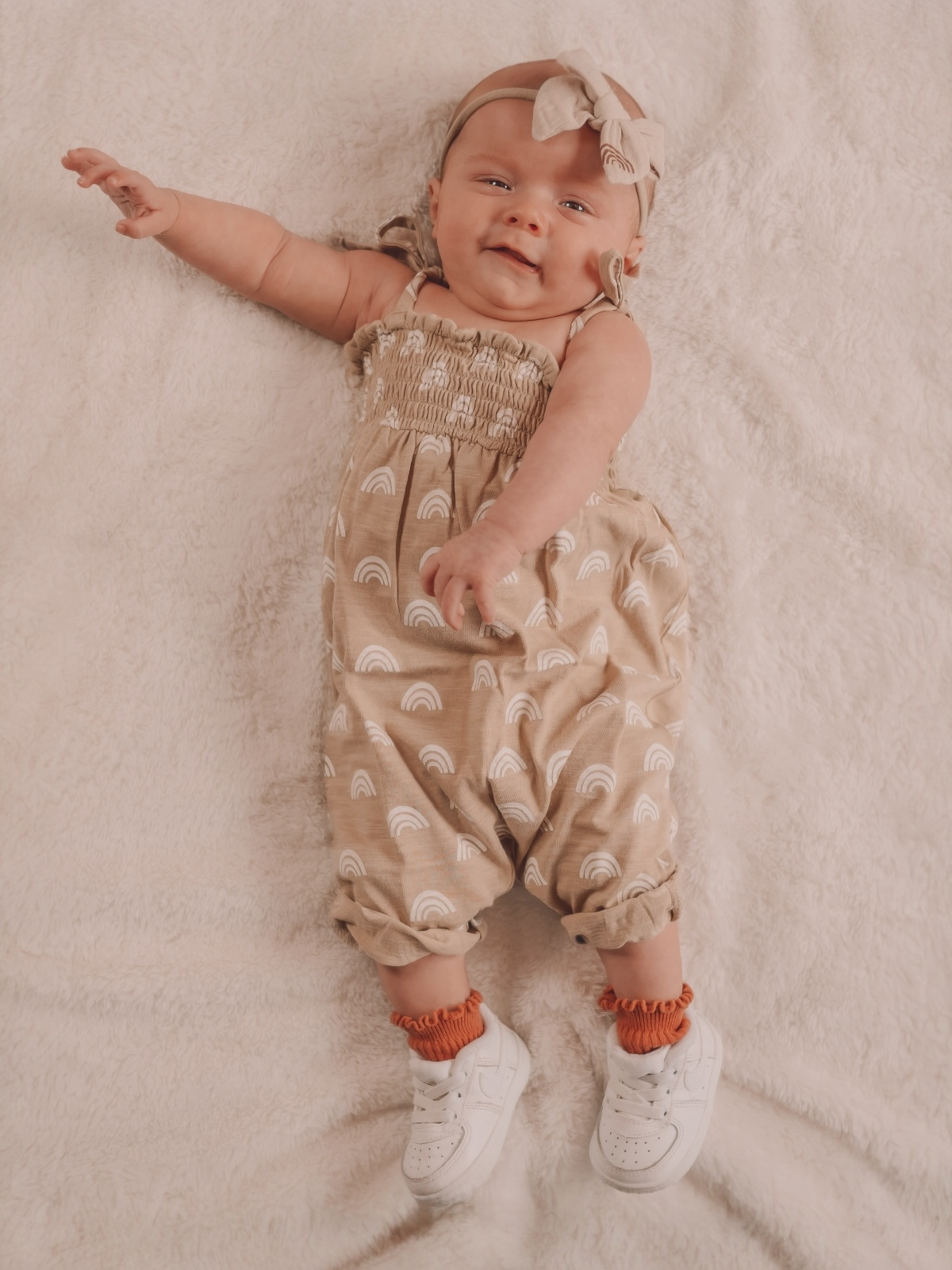 Smiling baby in a rainbow-patterned outfit and white sneakers, lying on a soft blanket.