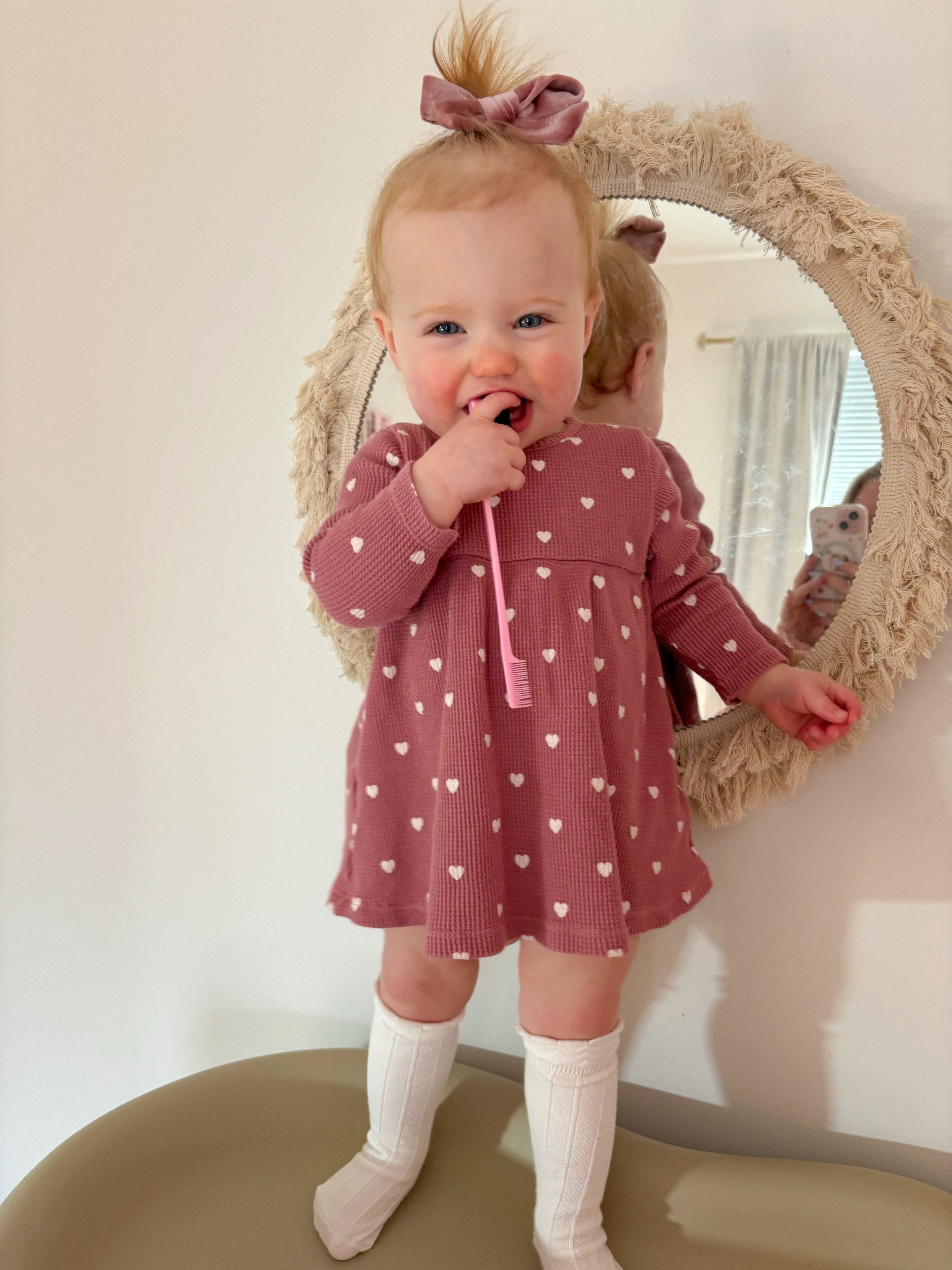 Smiling toddler in a heart-patterned dress holding a pink toothbrush, standing by a decorative mirror.