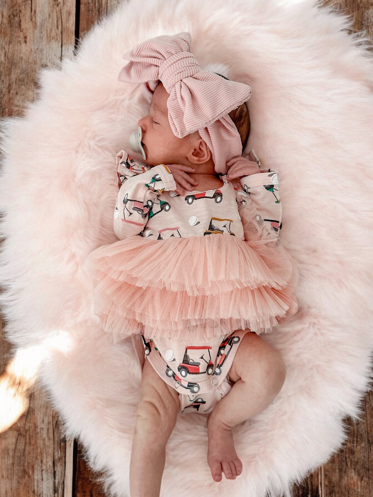Cute baby girl wearing a pink bow and a dress with cars, lying on a fluffy rug.