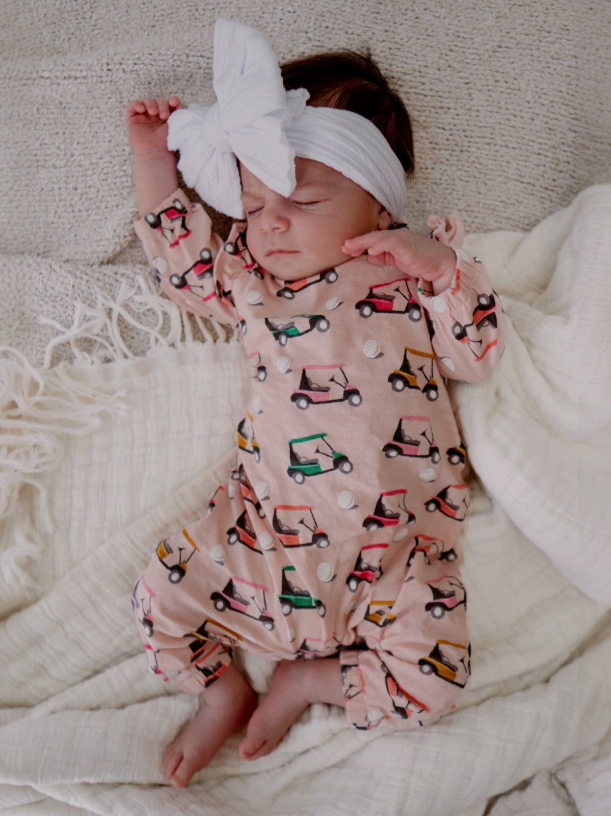 Sleeping baby in a patterned onesie with golf carts, wearing a large white bow headband on a soft blanket.