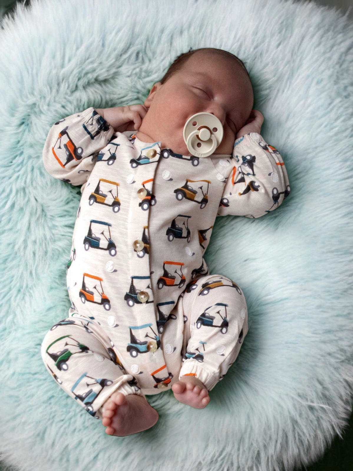 Infant sleeping on a fluffy blue blanket, wearing a golf cart-patterned onesie with a pacifier.