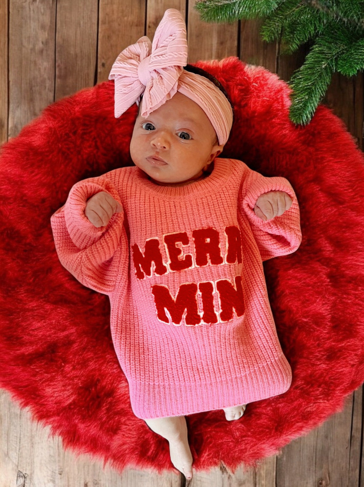 Baby wearing a pink "Merry" sweater, lying on a red fluffy blanket, with a matching pink headband and a wooden floor background.