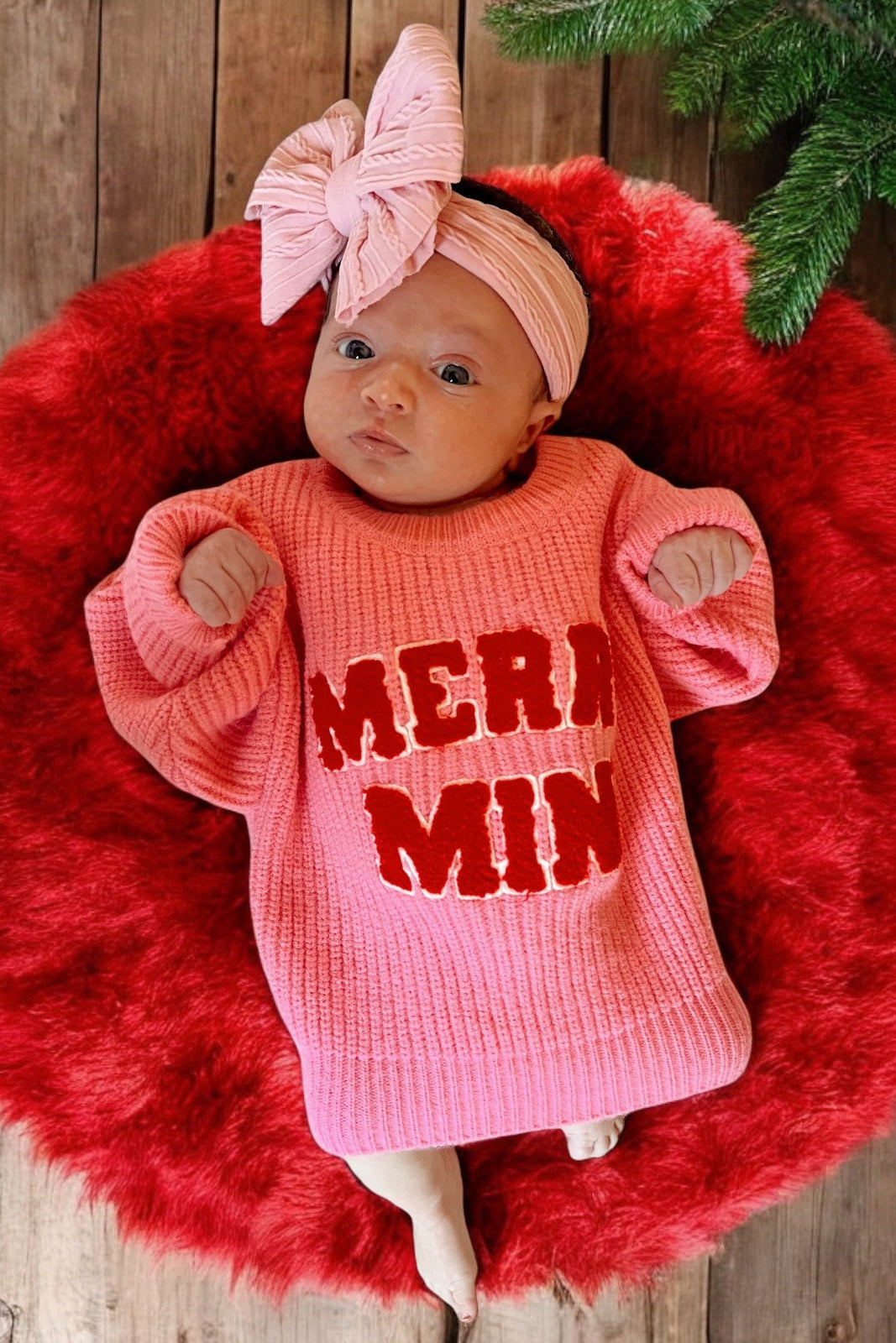 Baby wearing a pink "Merry" sweater, lying on a red fluffy blanket, with a matching pink headband and a wooden floor background.