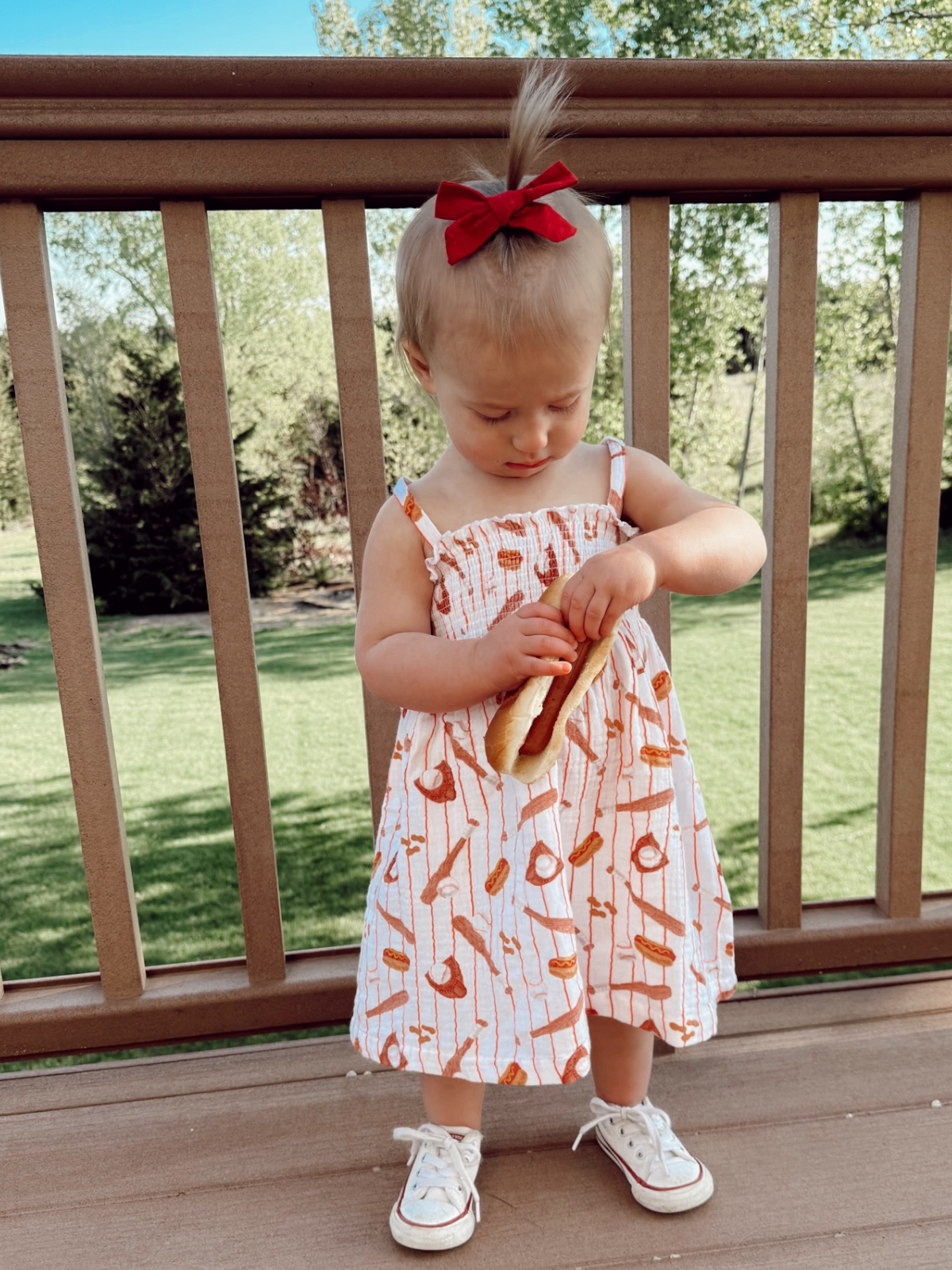 Toddler in a patterned dress and red bow holding a hot dog on a deck with green grass in the background.