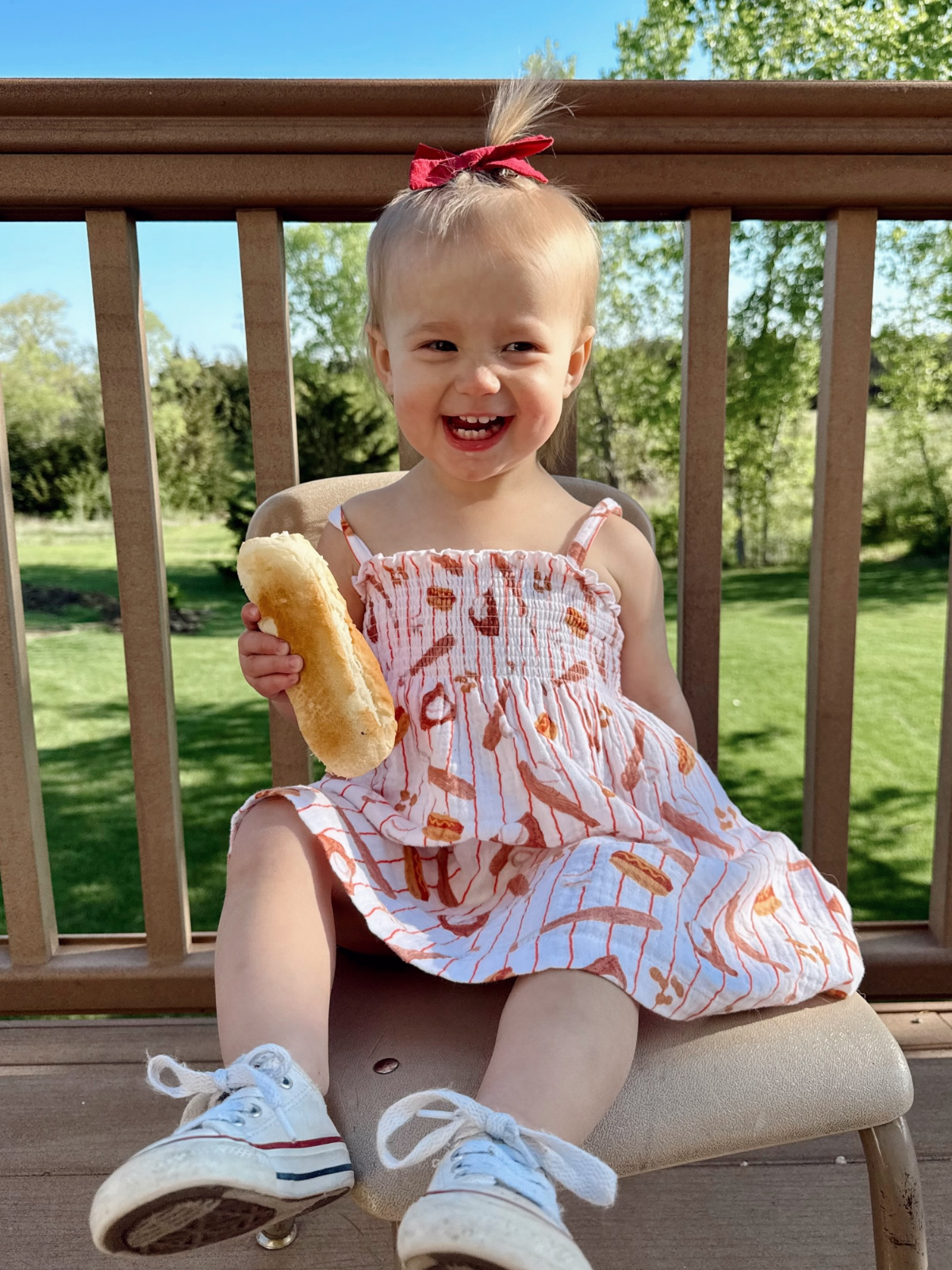 Smiling toddler in a striped dress holds a bun, sitting on a chair on a sunny deck with greenery in the background.