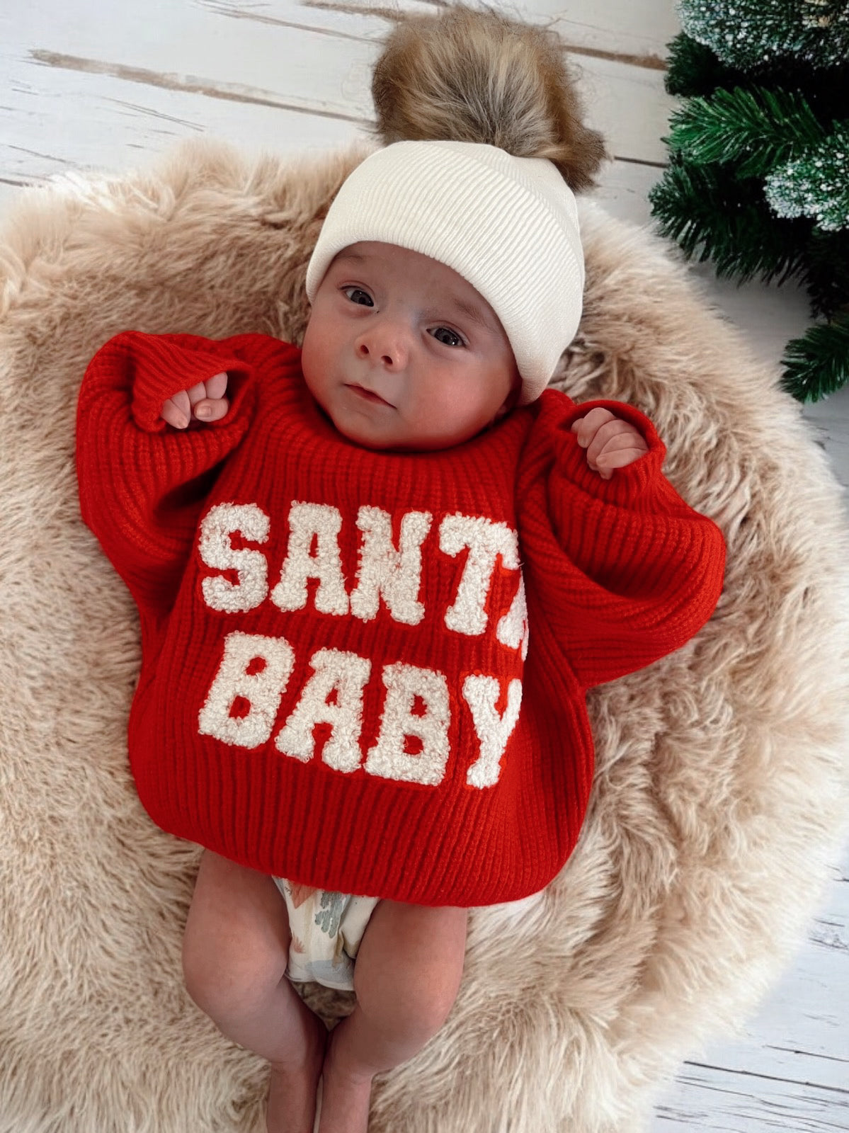 Baby wearing a red sweater with "SANTA BABY" text, lying on a fluffy rug with a festive background.