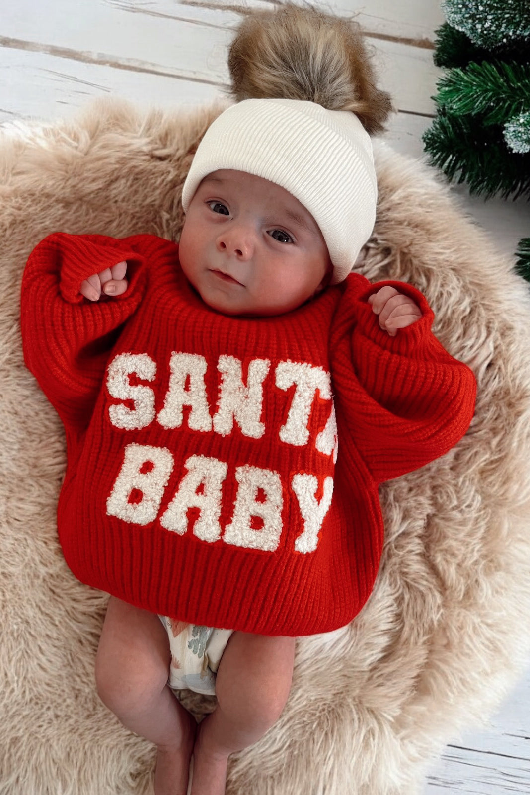 Baby wearing a red sweater with "SANTA BABY" text, lying on a fluffy rug with a festive background.