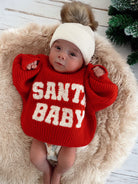 Baby wearing a red sweater with "SANTA BABY" text, lying on a fluffy rug with a festive background.