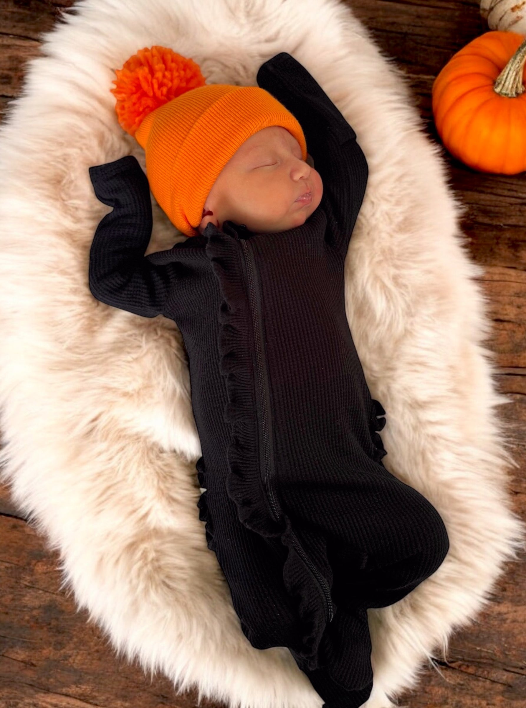 Baby sleeping peacefully on a soft fur blanket, wearing a black outfit and an orange pom-pom hat.