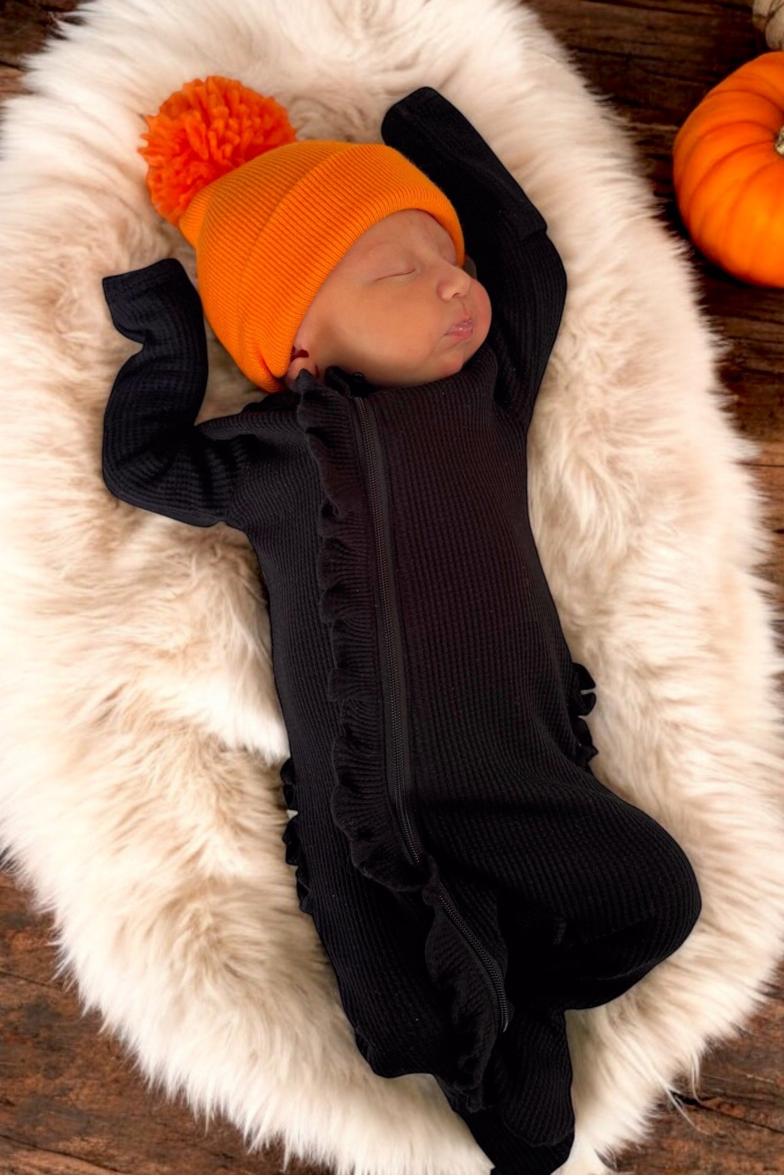 Baby sleeping peacefully on a soft fur blanket, wearing a black outfit and an orange pom-pom hat.