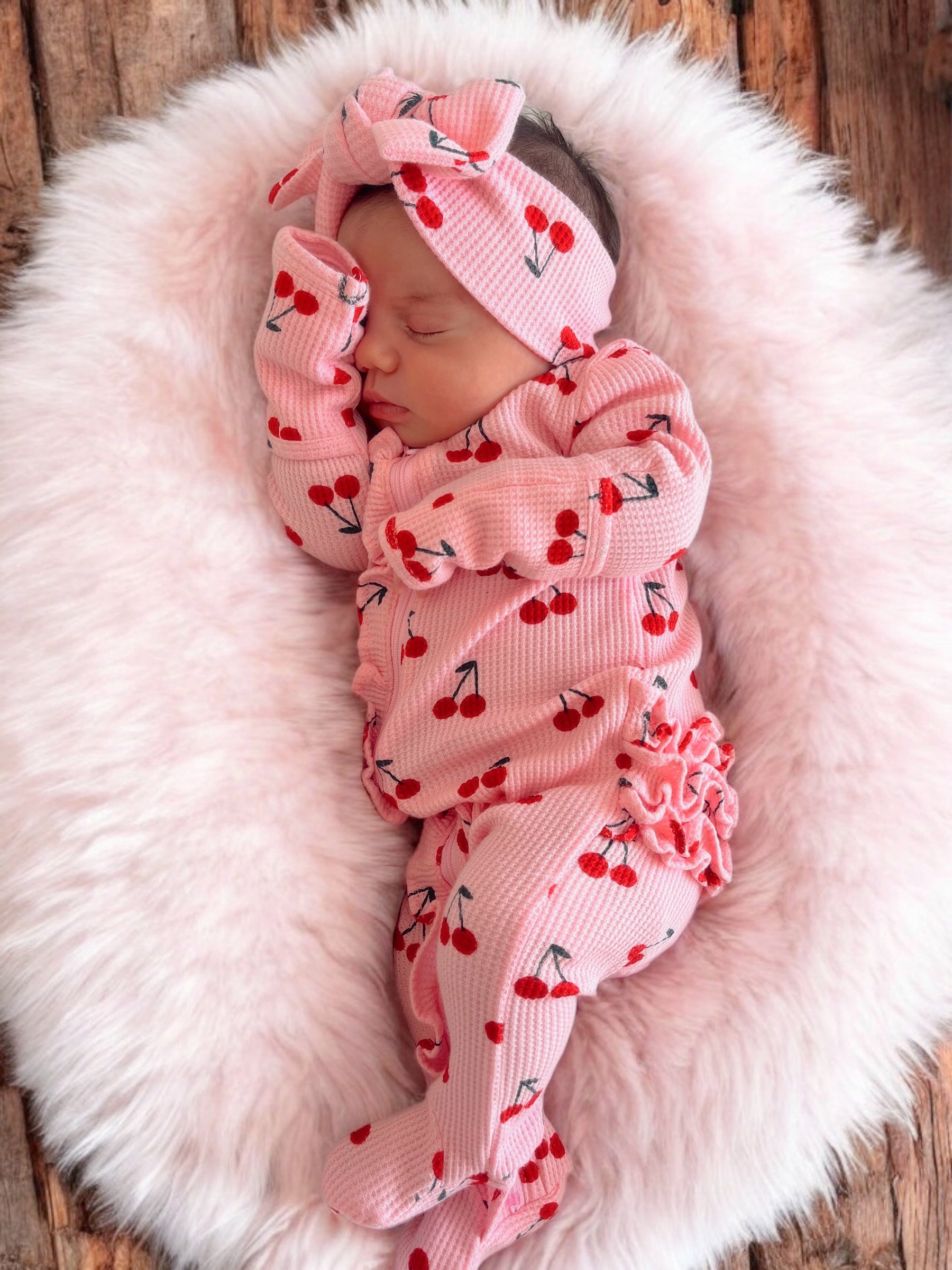 Baby girl peacefully sleeping in pink cherry-patterned outfit on fluffy blanket. Soft textures create a cozy atmosphere.