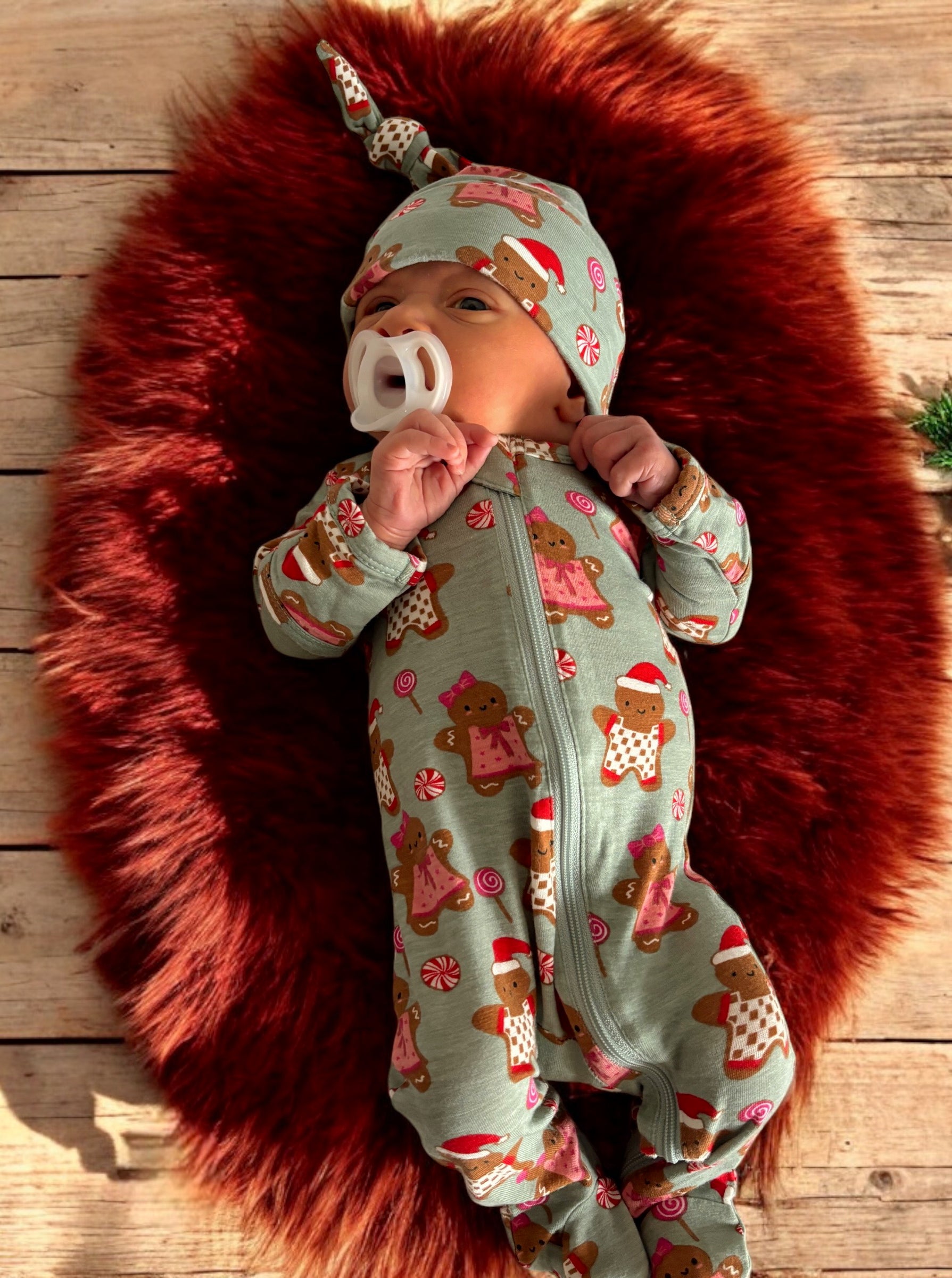 Baby in gingerbread-themed onesie and hat, lying on a fluffy red rug with a wooden background.