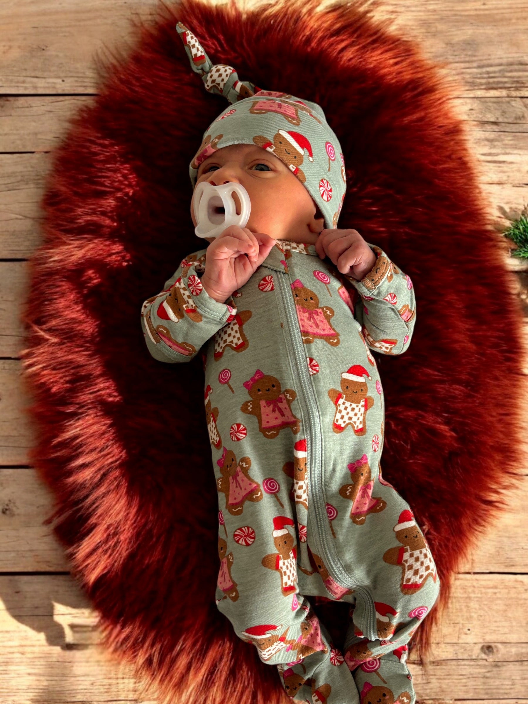 Baby in gingerbread-themed onesie and hat, lying on a fluffy red rug with a wooden background.