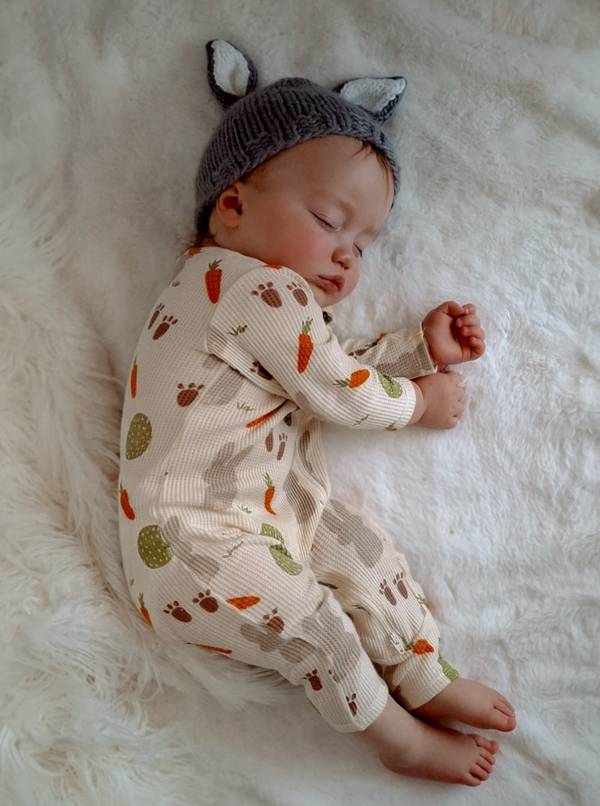 Baby sleeping peacefully on a soft blanket, wearing a patterned onesie and a cute animal ear hat.