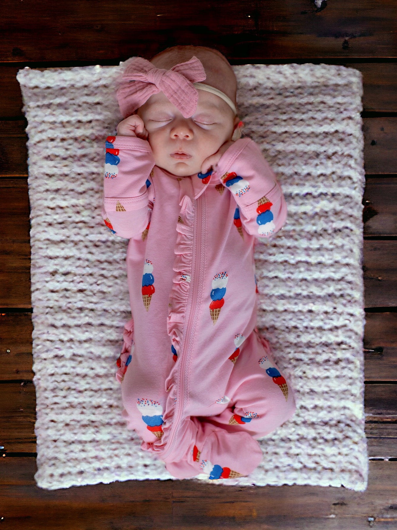 Sleeping baby in pink ice cream-themed outfit, resting on a soft blanket on wooden floor.