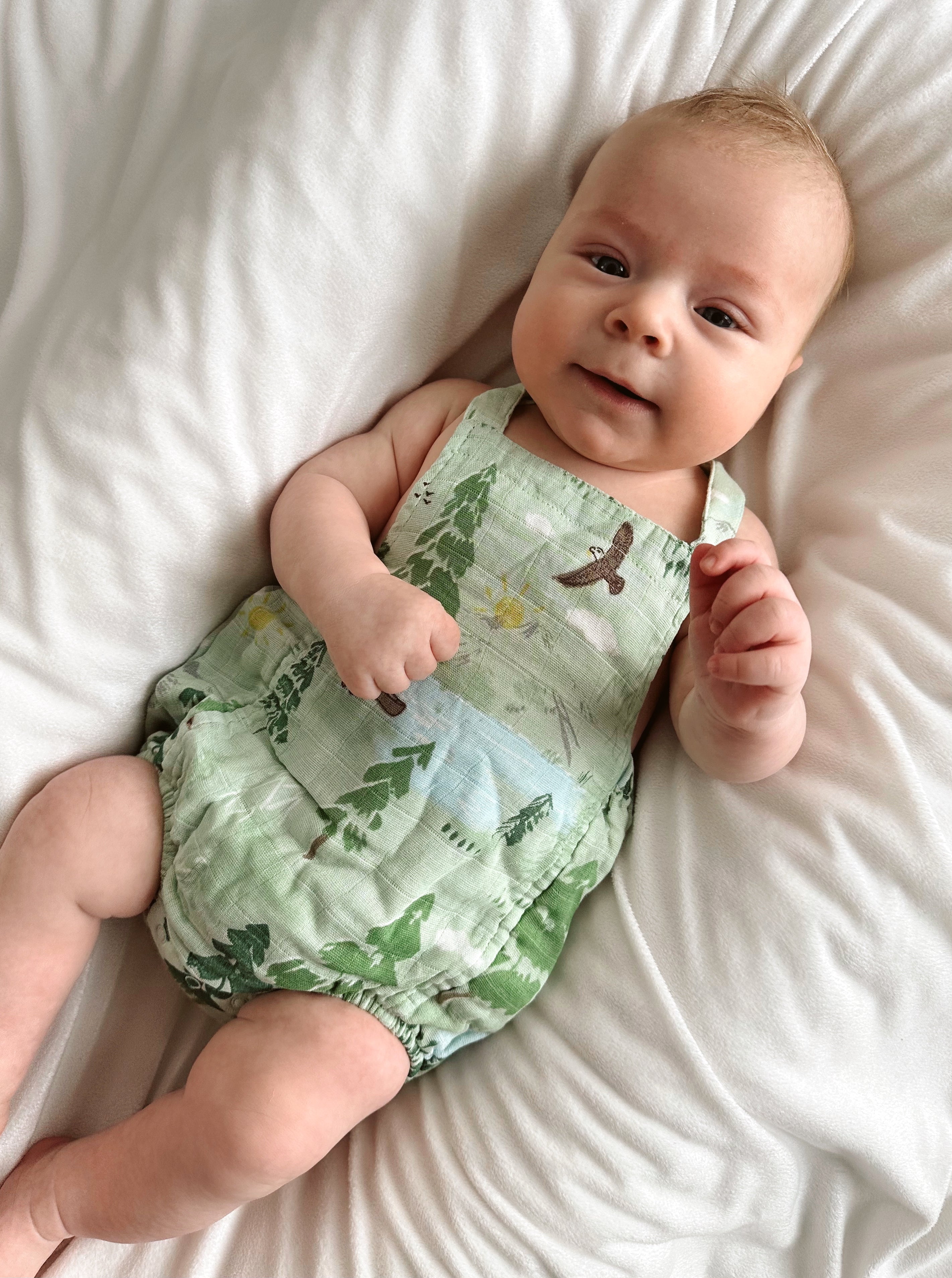 Smiling baby in a green patterned onesie, seated on a white blanket, with hands raised and playful expression.