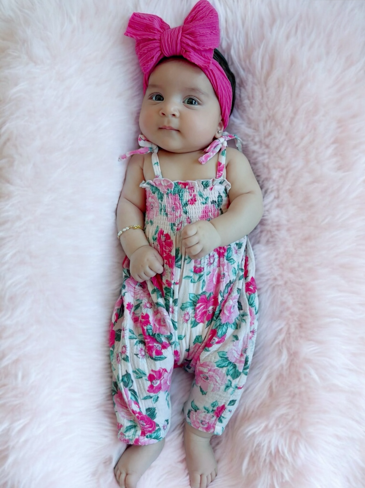 Baby girl in floral outfit and pink headband, smiling on a soft pink blanket.