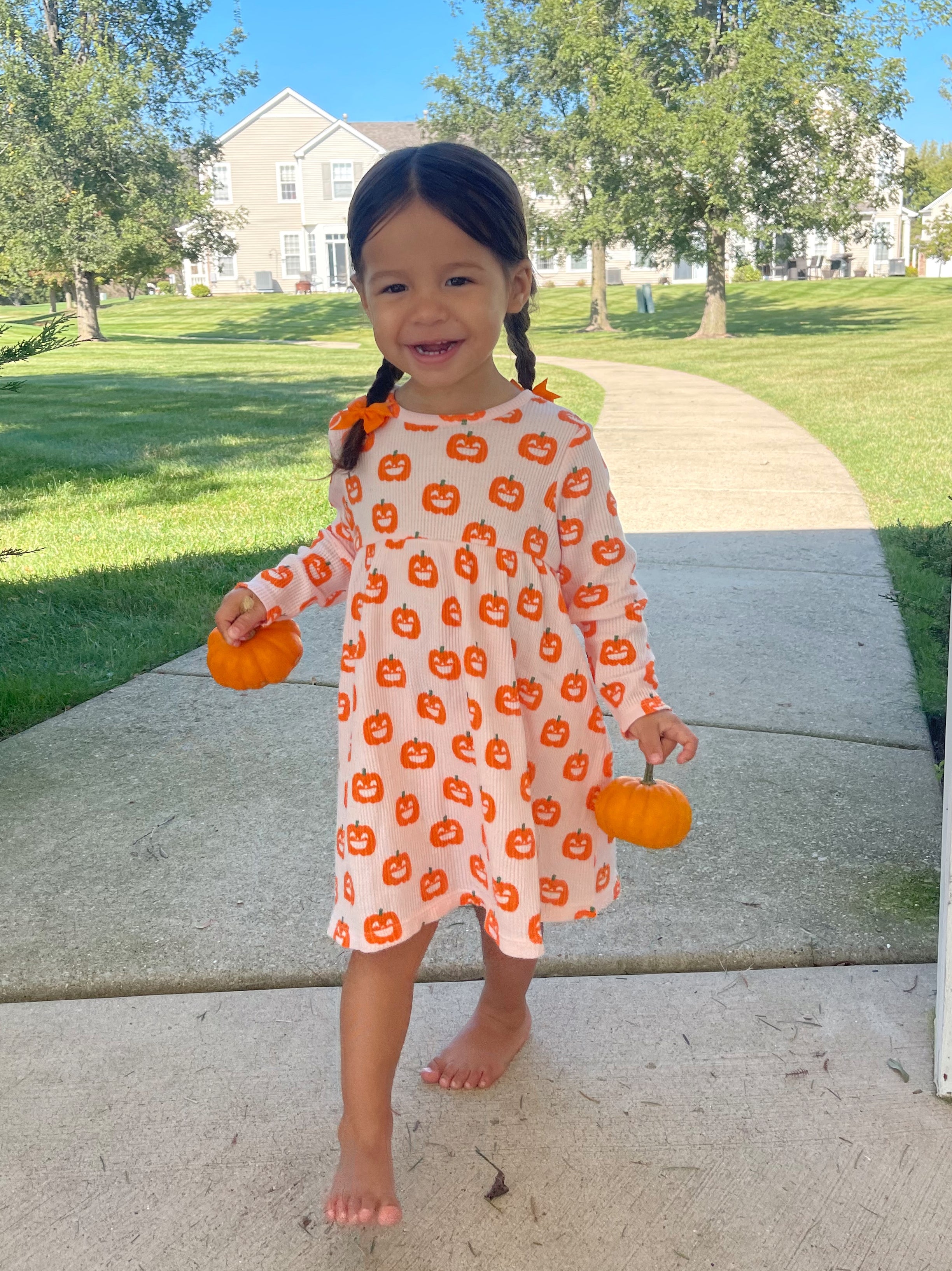 Smiling child in pumpkin-patterned dress, holding mini pumpkins outdoors on a sunny day.