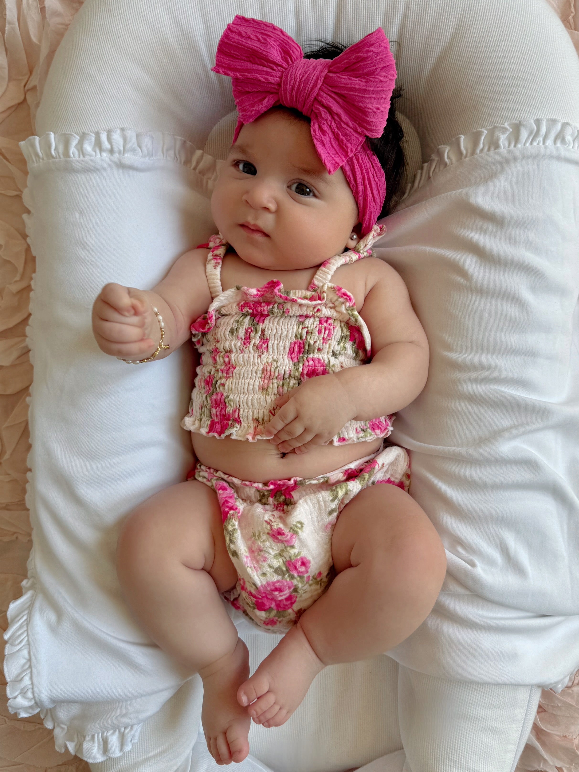 Baby girl with a pink bow, wearing a floral outfit, lying on a soft pillow.