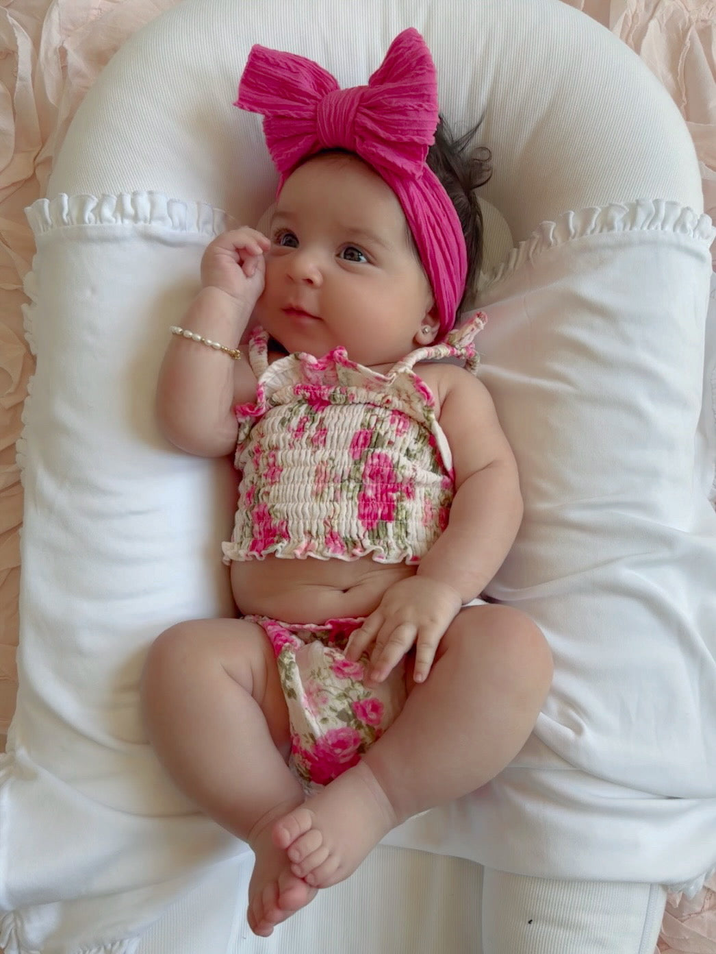 Baby girl in floral outfit with pink headband, sitting on a white pillow against a light pink background.