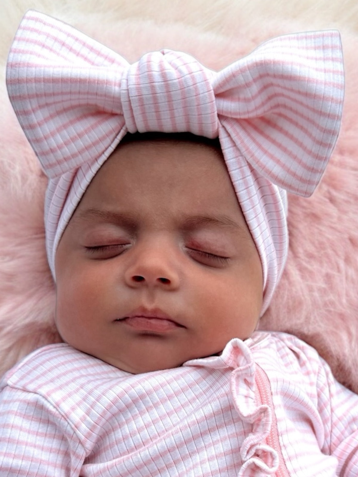 Sleeping baby girl in a pink and white striped headband, resting on a soft, fluffy surface.