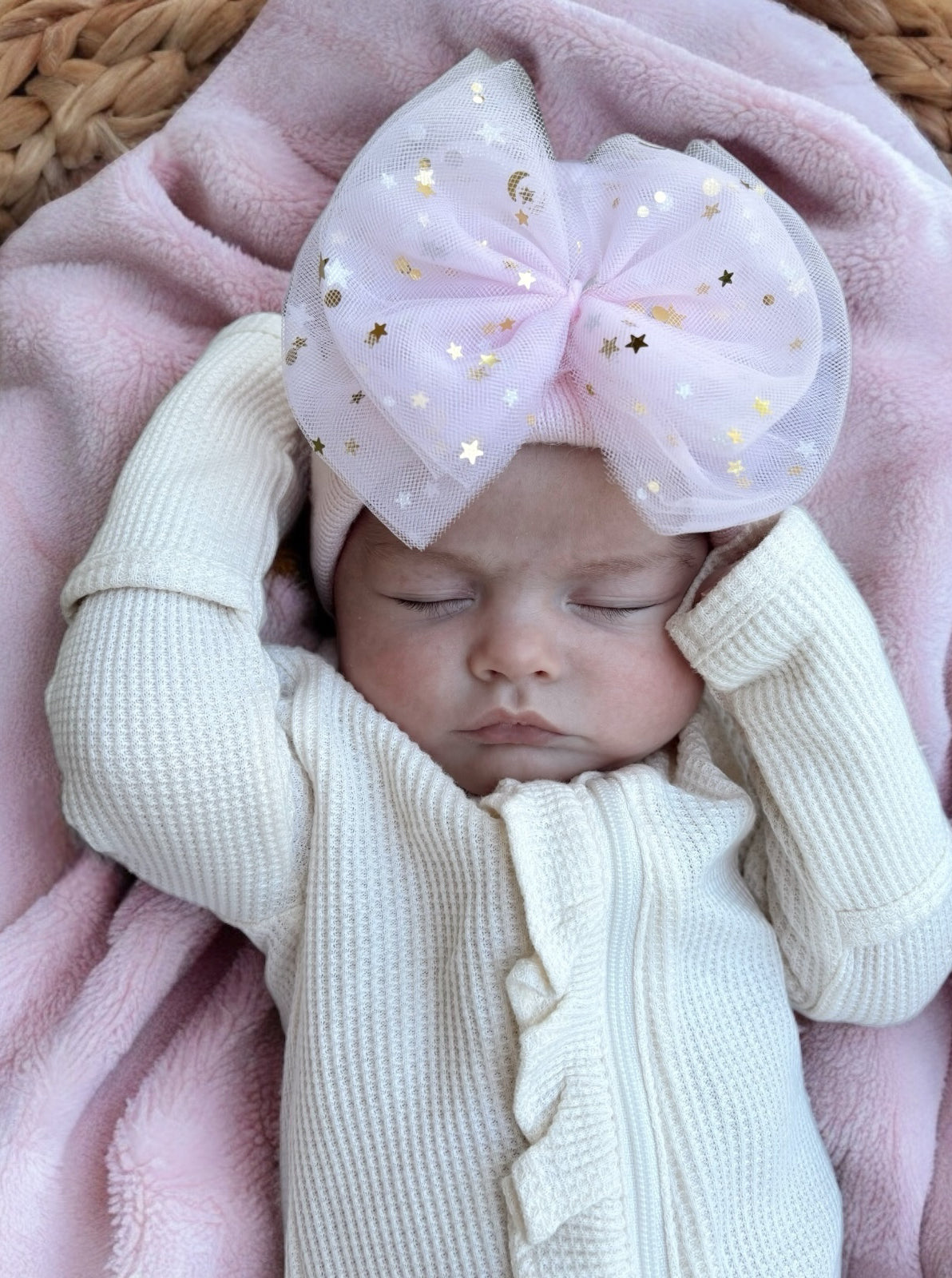 Baby girl sleeping with a large pink bow and white outfit, lying on a pink blanket.